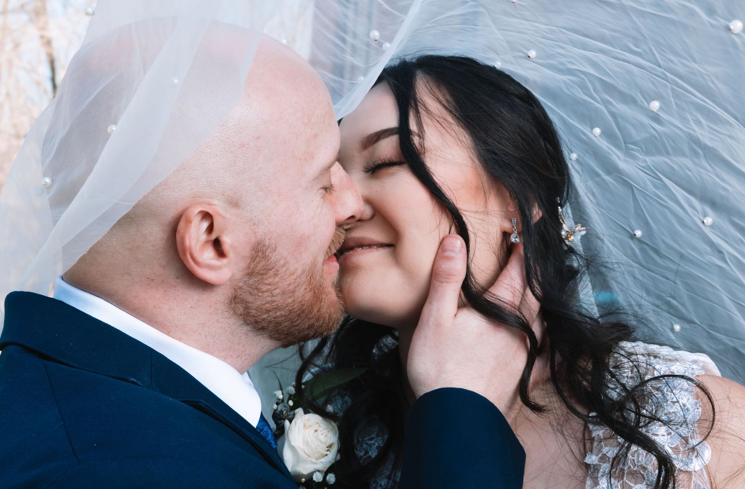 Groom gently holds brides face as they share a close romantic kiss beneath a wedding veil.