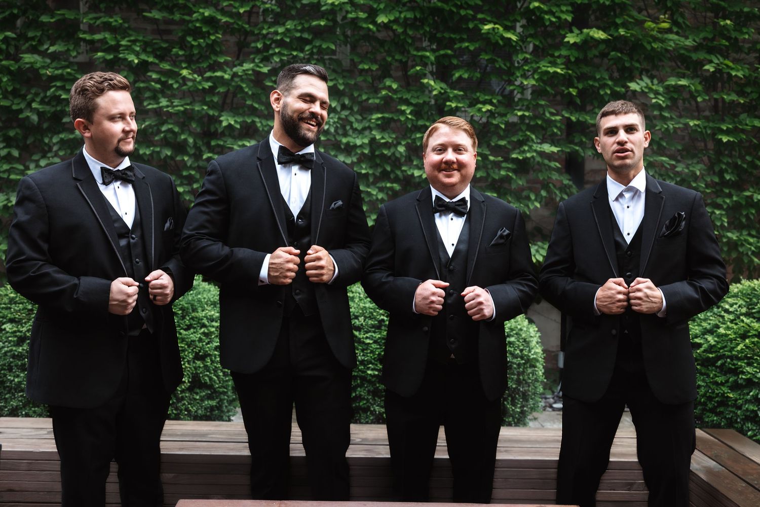 Four groomsmen laughing and adjusting their black tuxedo jackets outdoors against a lush green ivy wall.