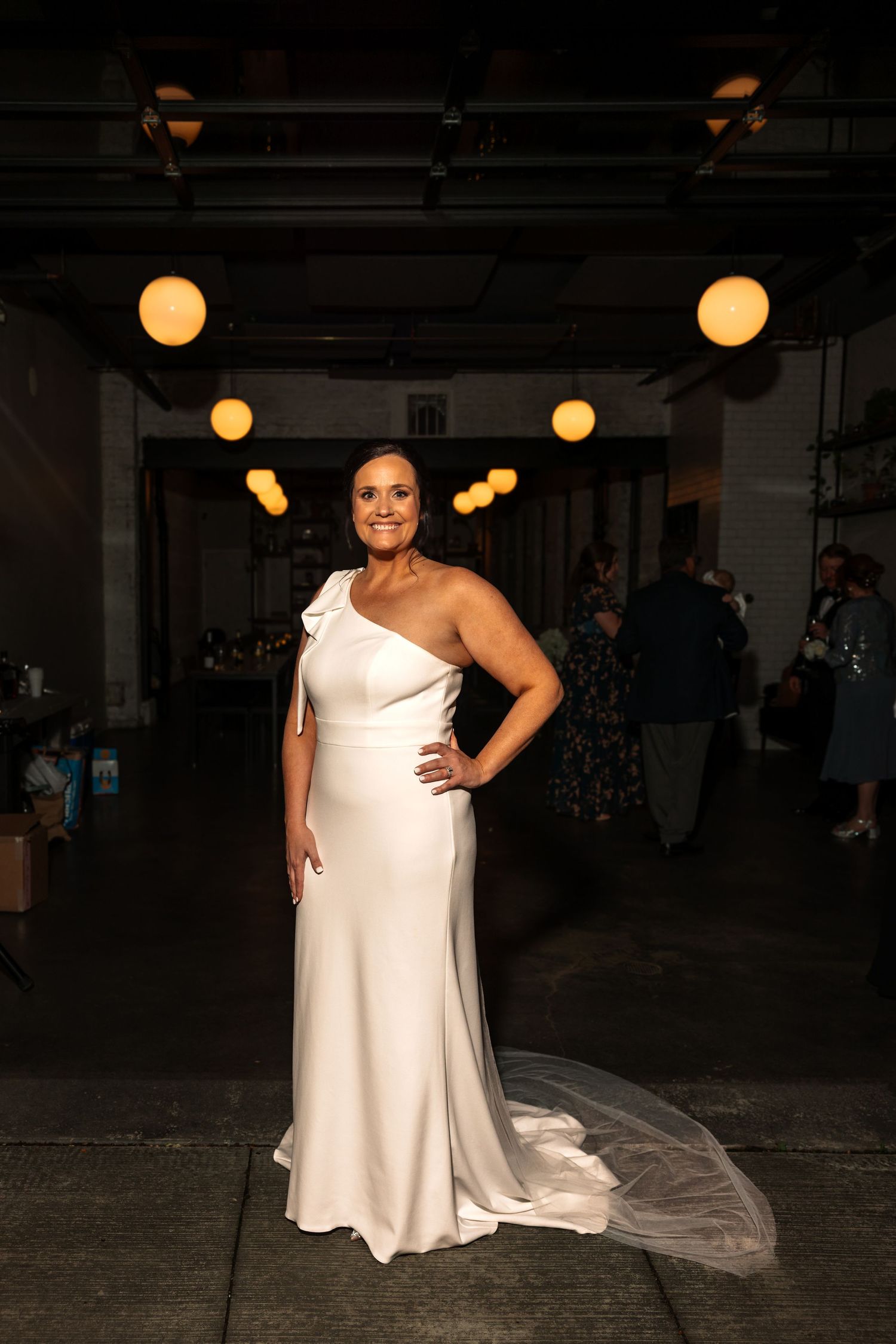 Radiant bride in fitted one-shoulder white wedding dress with train stands confidently in warmly lit industrial venue.