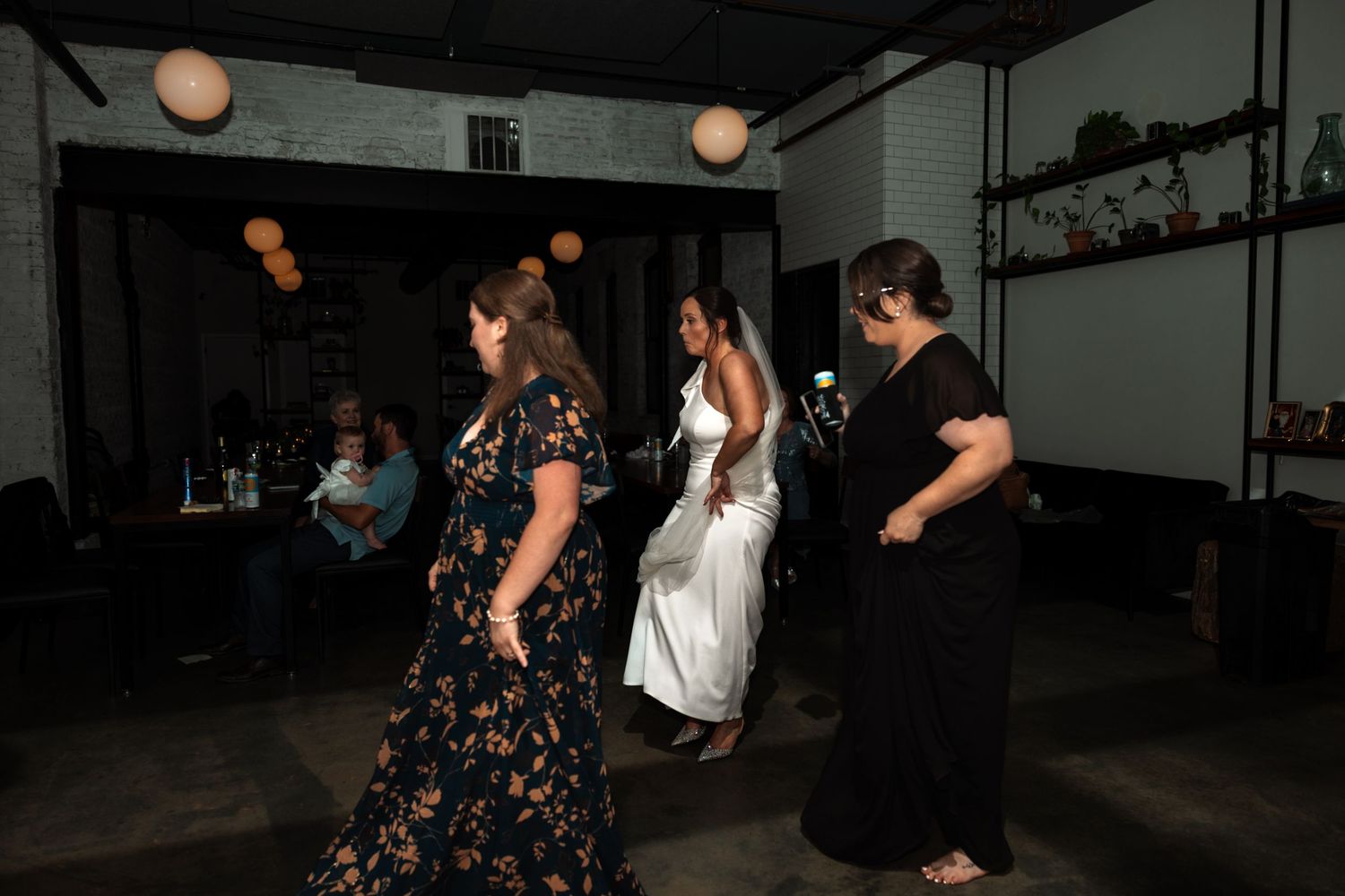 Bride in white gown dancing with guests in floral and black dresses at a wedding reception.