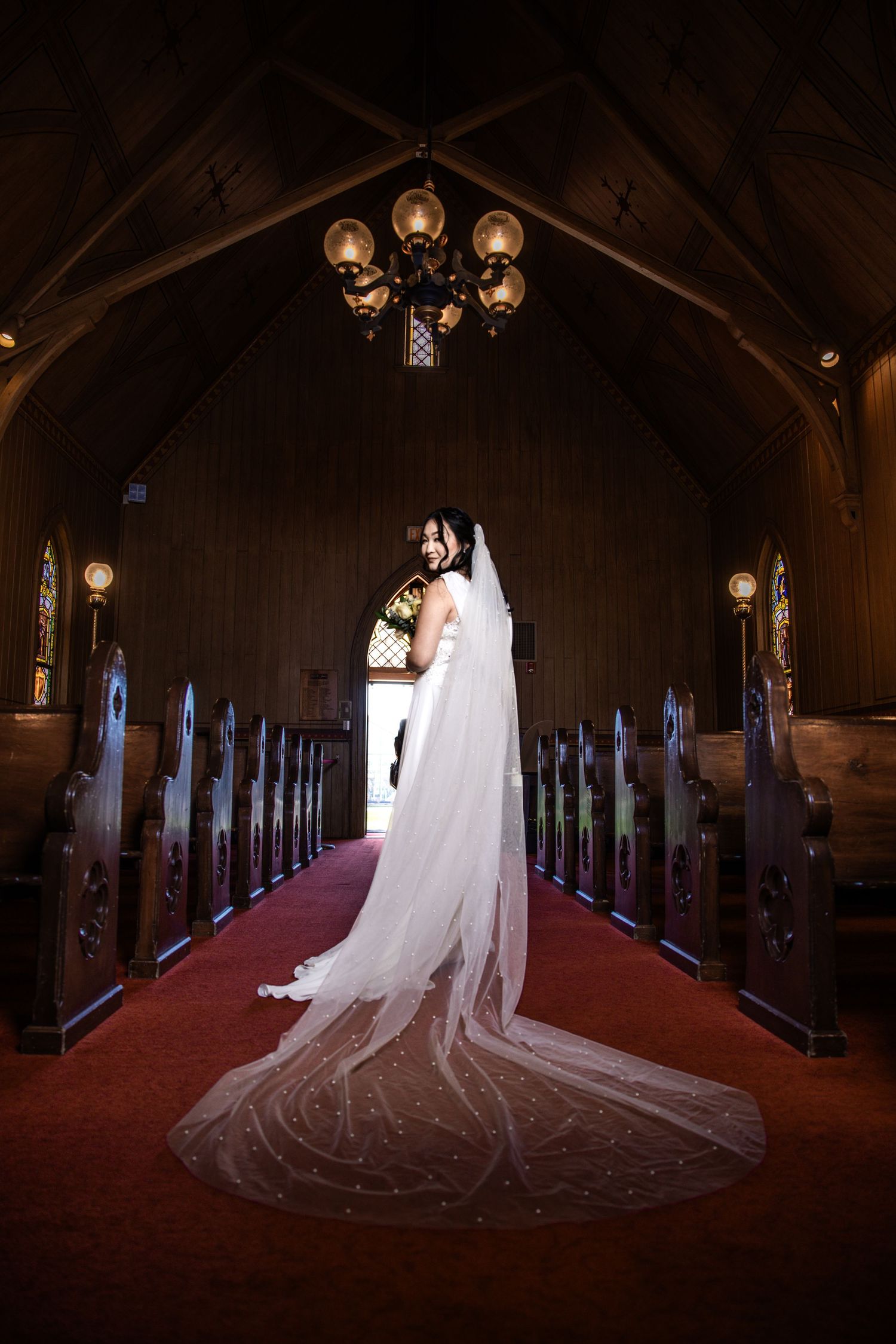 Bride in a stunning white gown with an elegant cathedral veil stands alone in a beautiful wooden church aisle.