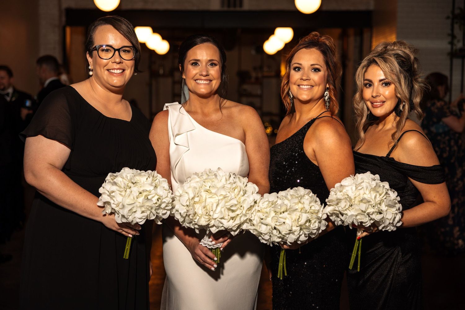 Bride in asymmetric white gown with bridesmaids in black holding white hydrangea bouquets at reception.