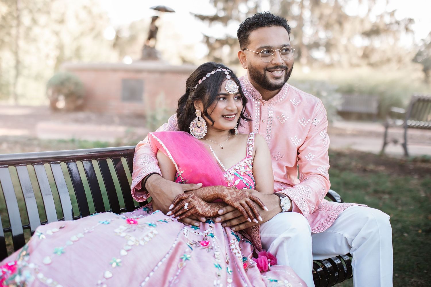 Indian bride and groom in pink traditional attire sitting together on outdoor bench.