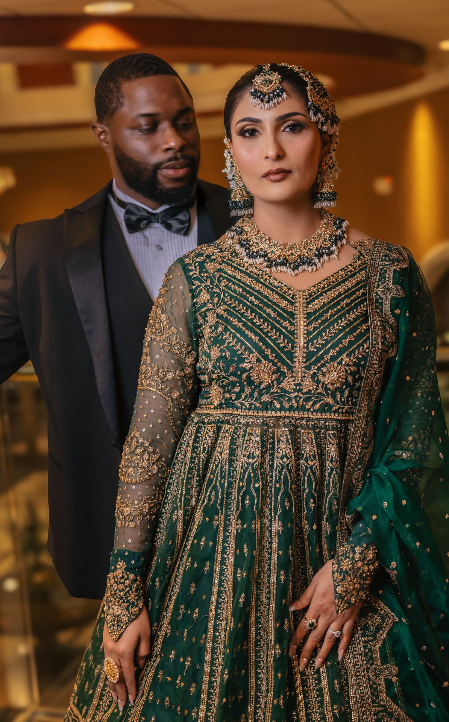 Bride in stunning emerald green embroidered lehenga with gold jewelry stands with groom in black tuxedo.