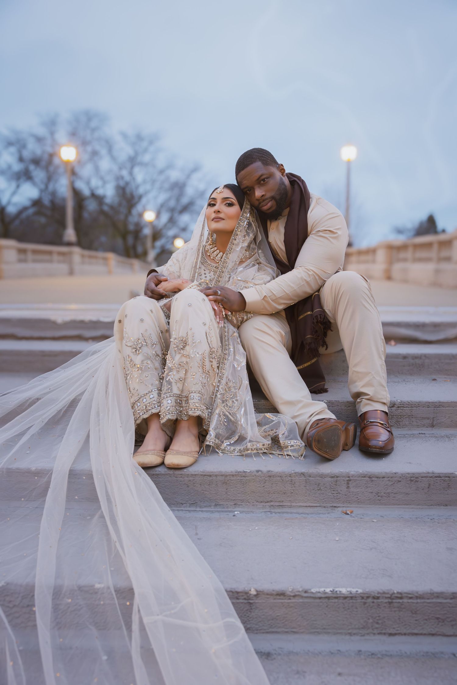 South Asian bride and groom in traditional cream wedding attire sit together on outdoor stone steps.