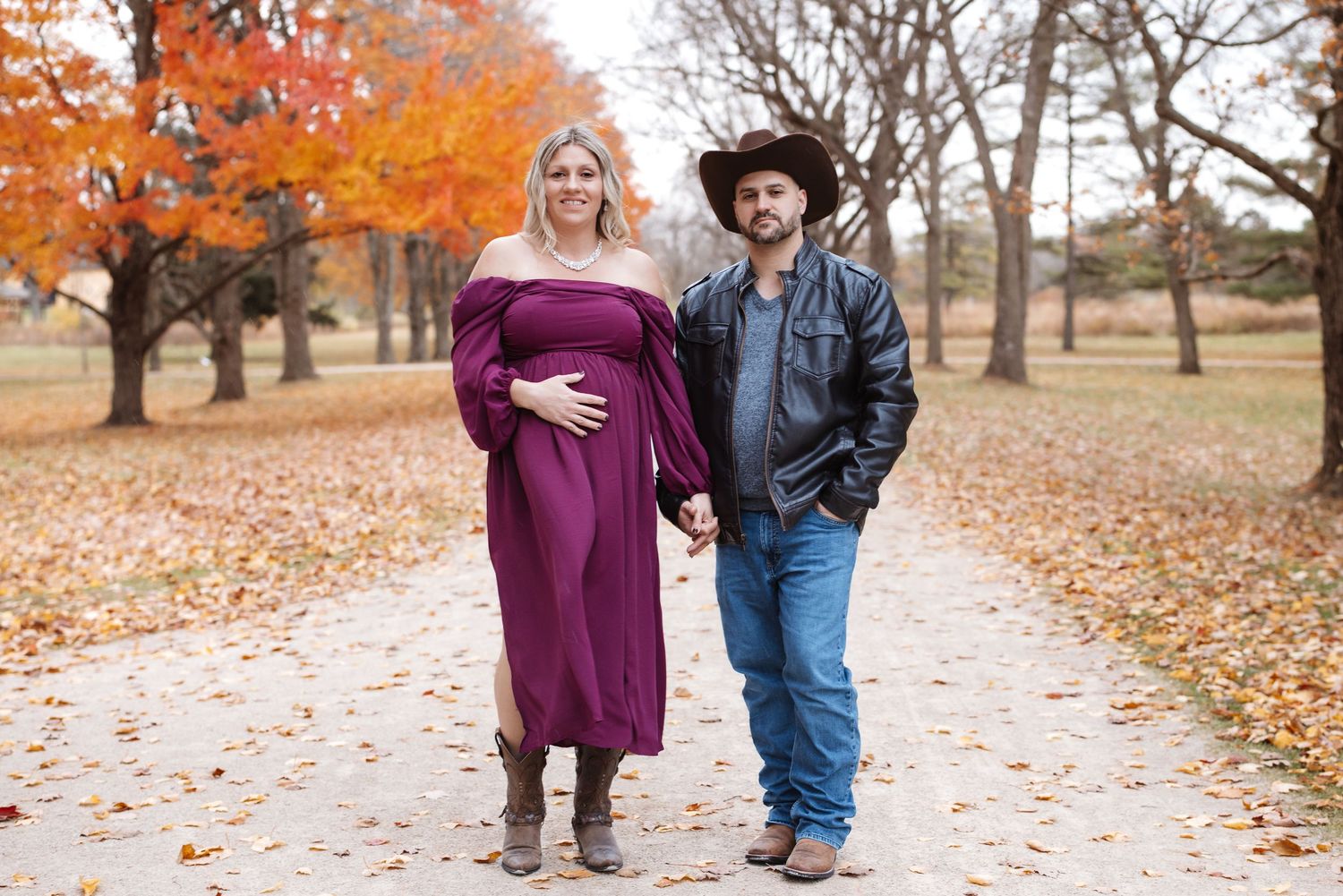 Pregnant couple holding hands on leaf-covered path surrounded by vibrant fall foliage.