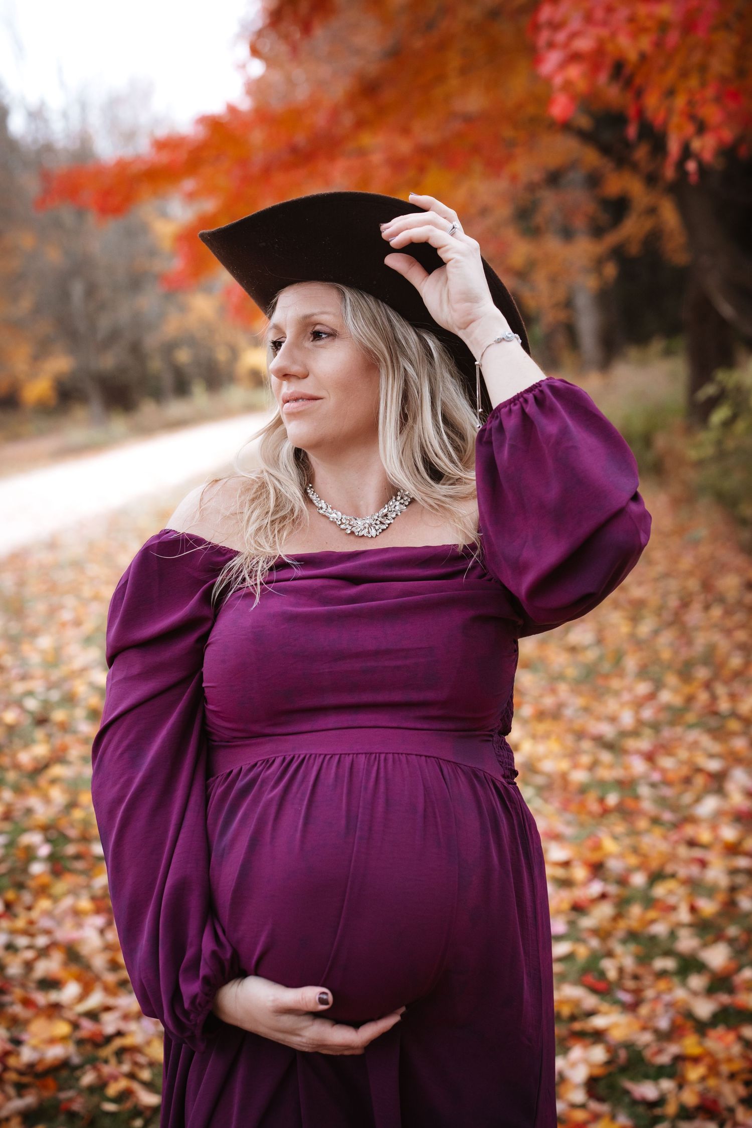 Pregnant woman in purple dress wearing graduation cap among autumn foliage.