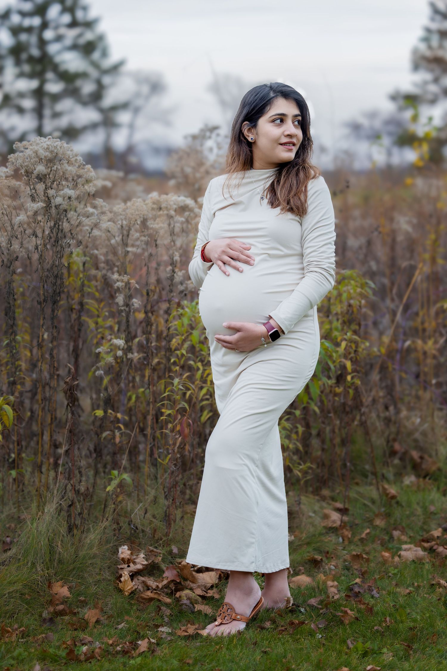 Pregnant woman in white dress cradling baby bump outdoors in autumn setting.