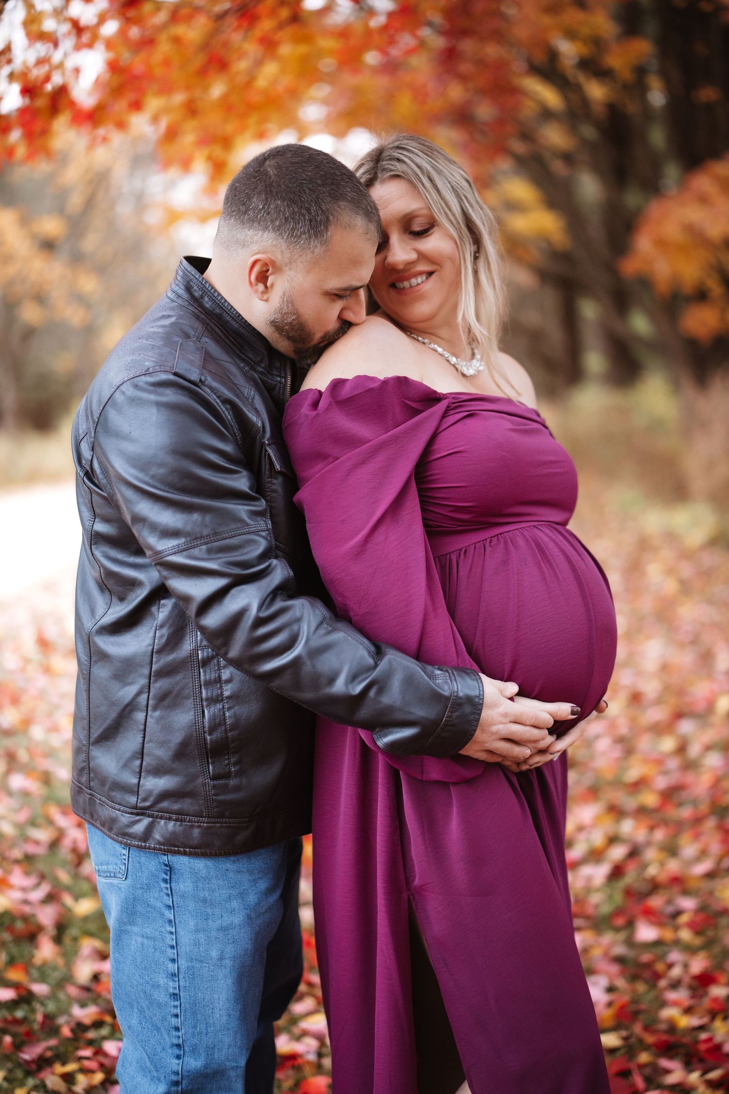 Couple sharing tender moment outdoors, man kissing pregnant woman in purple dress amid autumn leaves.