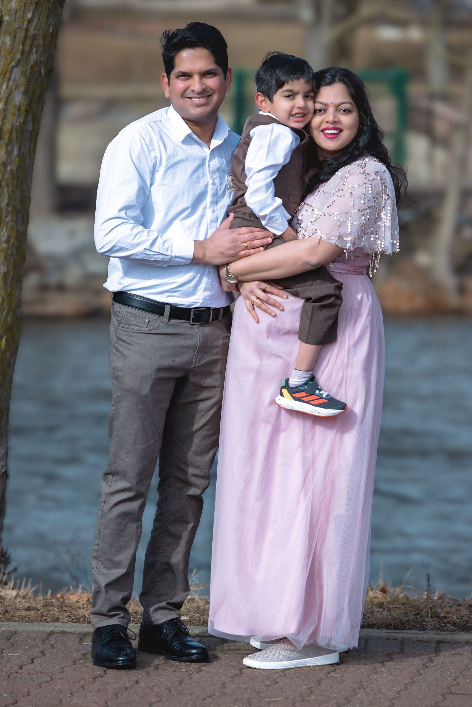 Smiling Indian family posing outdoors near water, father and pregnant mother in pink dress holding their young son.