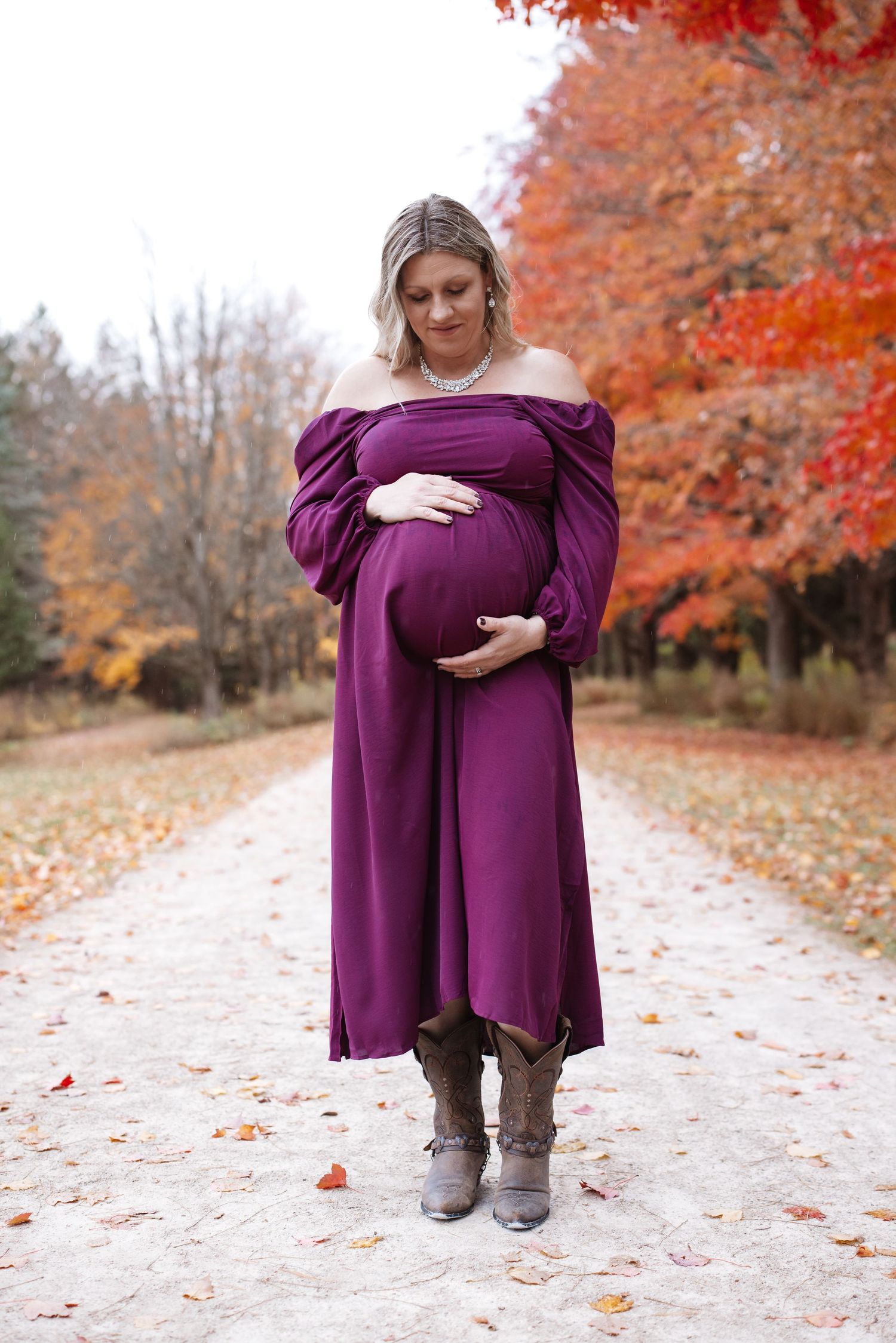 Pregnant woman in purple off-shoulder maternity dress and boots standing on scenic fall pathway.