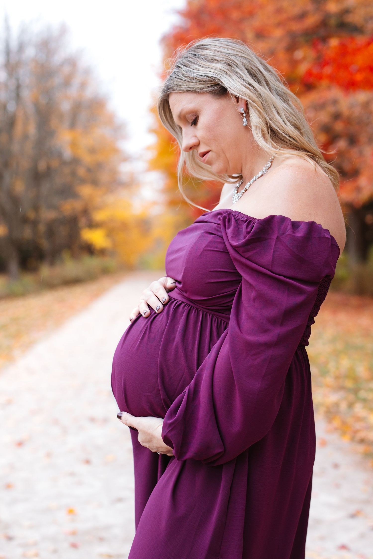 Pregnant woman in purple off-shoulder gown tenderly holding baby bump surrounded by fall foliage.