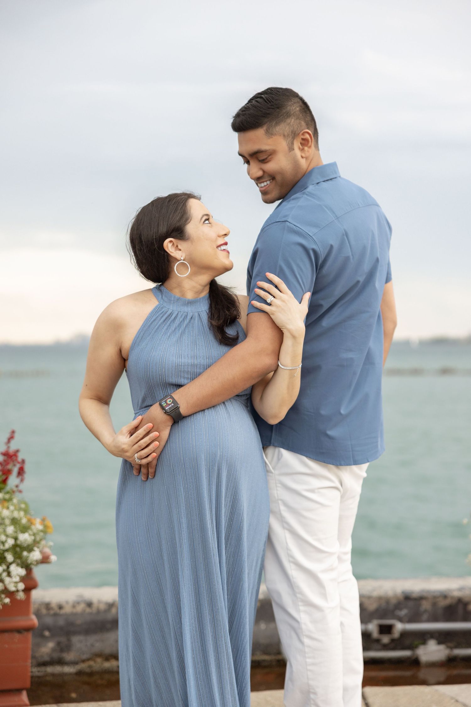 Smiling pregnant couple in matching blue outfits embracing by the waterfront.