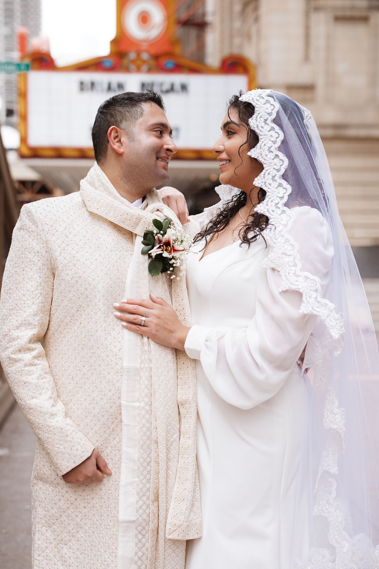 Newlyweds share a romantic glance outside the Chicago Theatre, groom in sherwani and bride in lace veil.