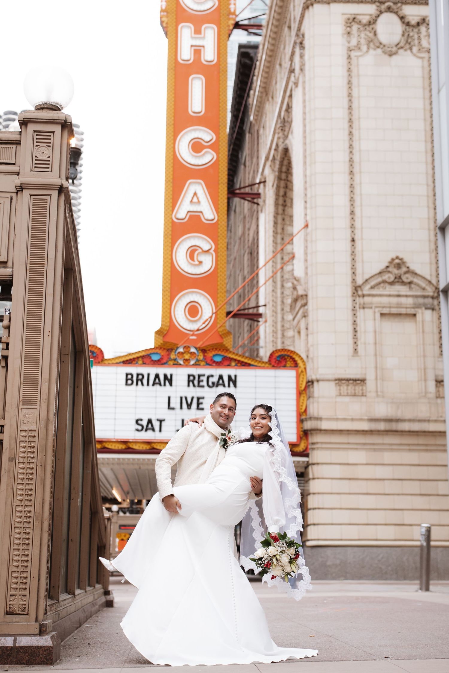 Newlyweds pose in front of Chicago Theatre sign with Brian Regan Live marquee below.