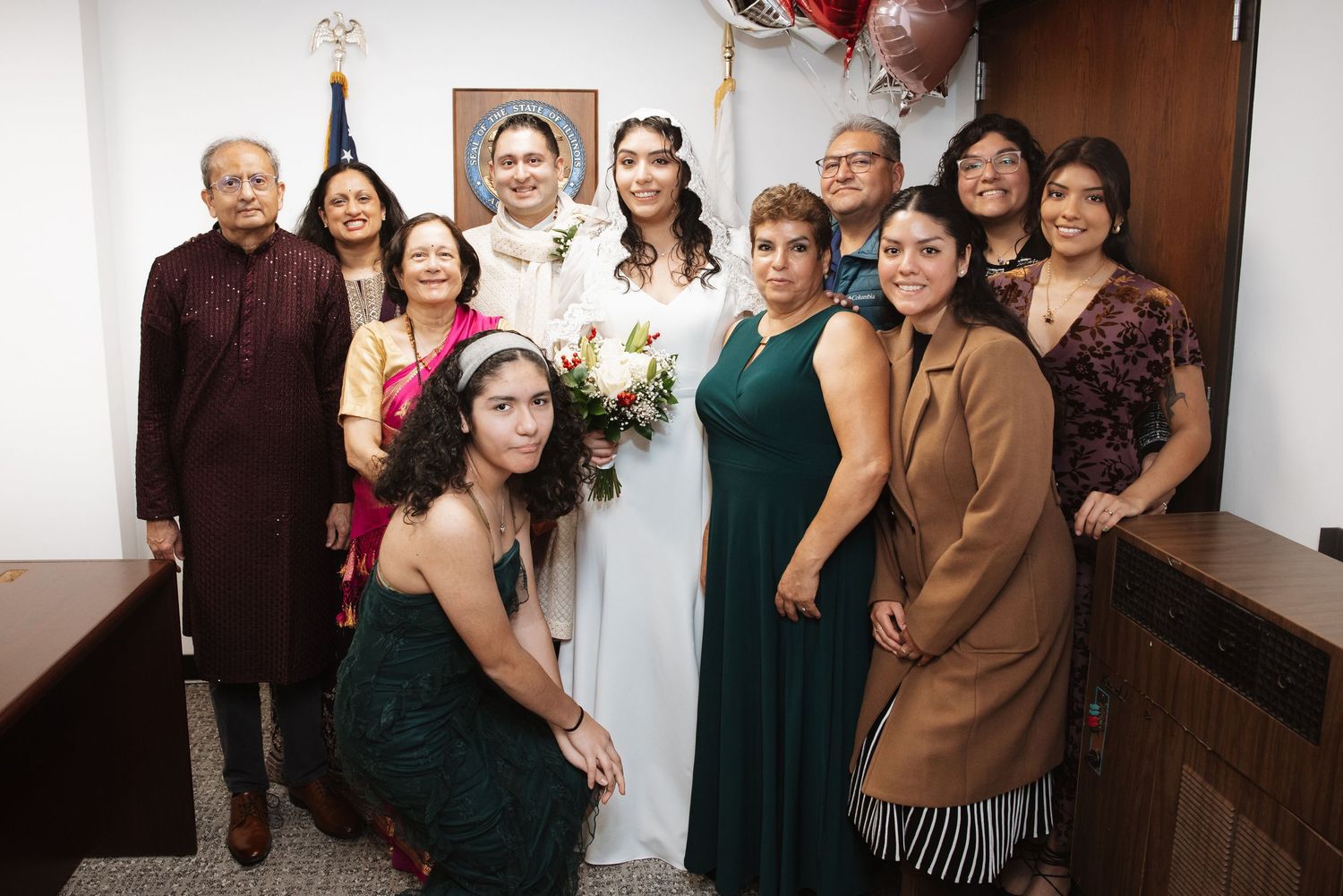 Wedding couple in white attire poses with large family group including elderly relatives in decorated ceremony room.
