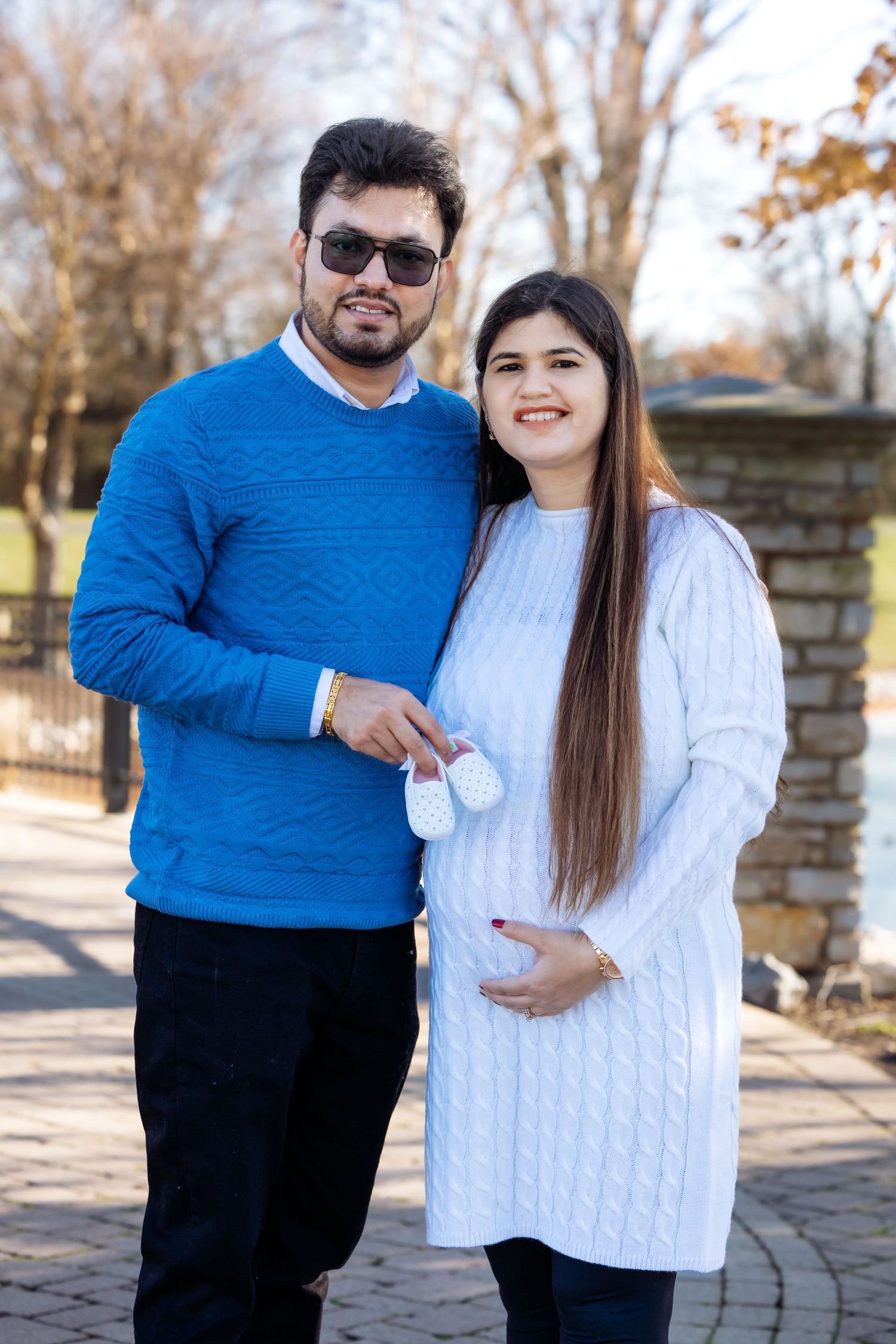 Happy couple announcing pregnancy outdoors, man in blue sweater holding baby booties beside pregnant woman in white dress.
