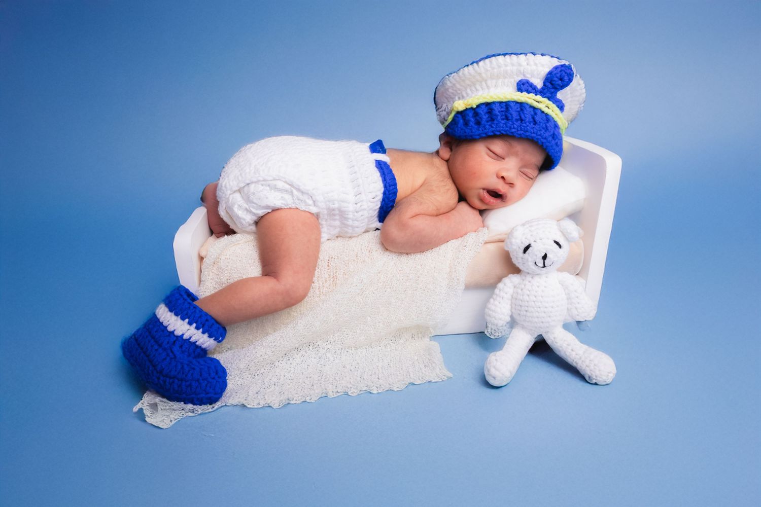 Sleeping newborn in white and blue sailor crochet outfit with teddy bear on blue background.