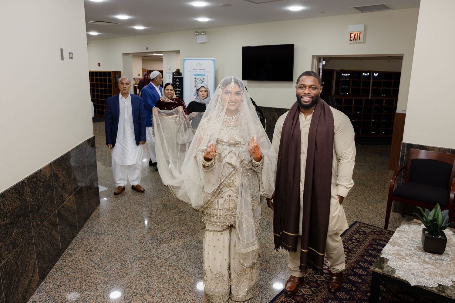 A bride in white South Asian bridal attire walks alongside a man in traditional brown dress inside a mosque lobby.