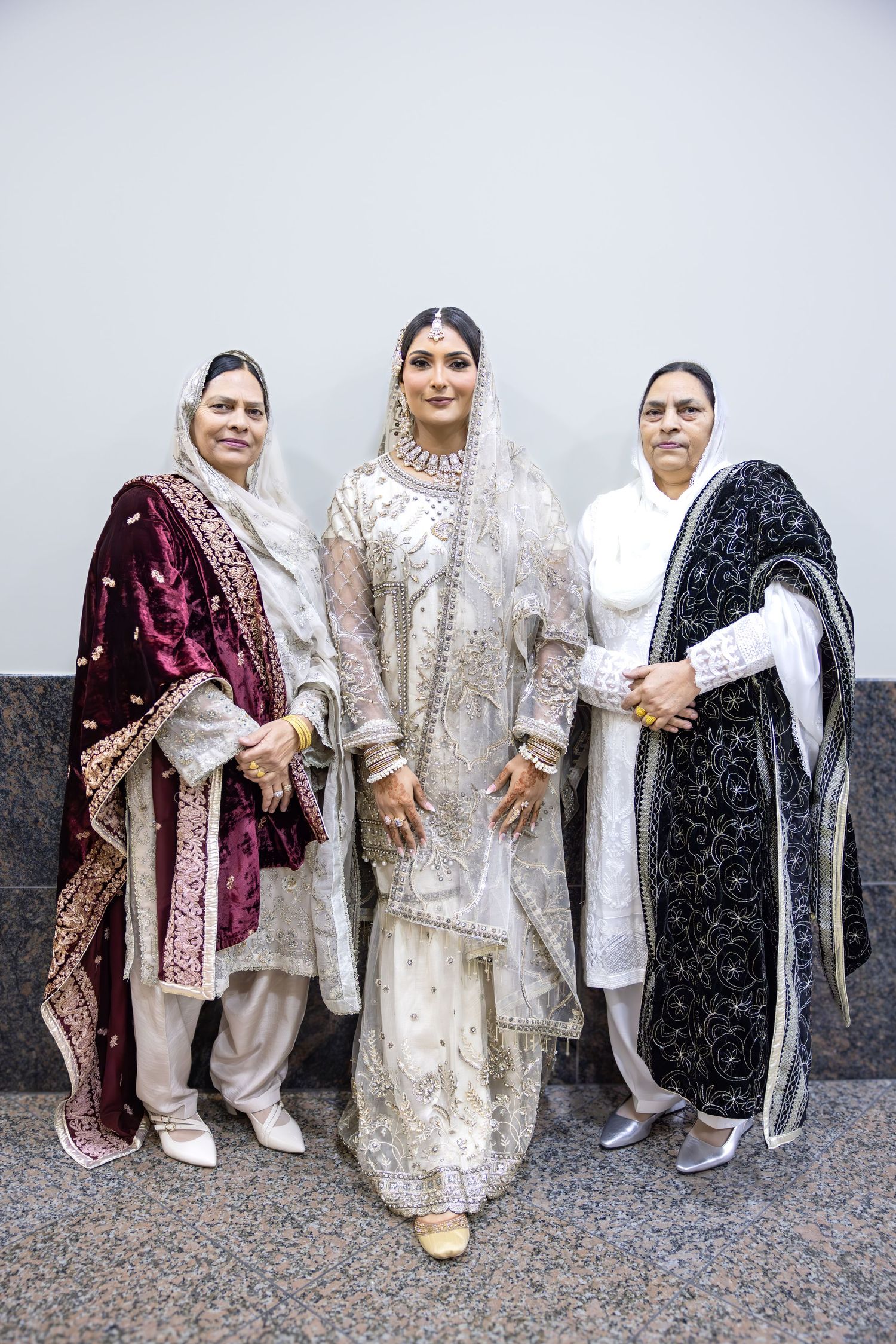 A bride in white embroidered bridal attire stands between two older women in traditional South Asian dress and headscarves.