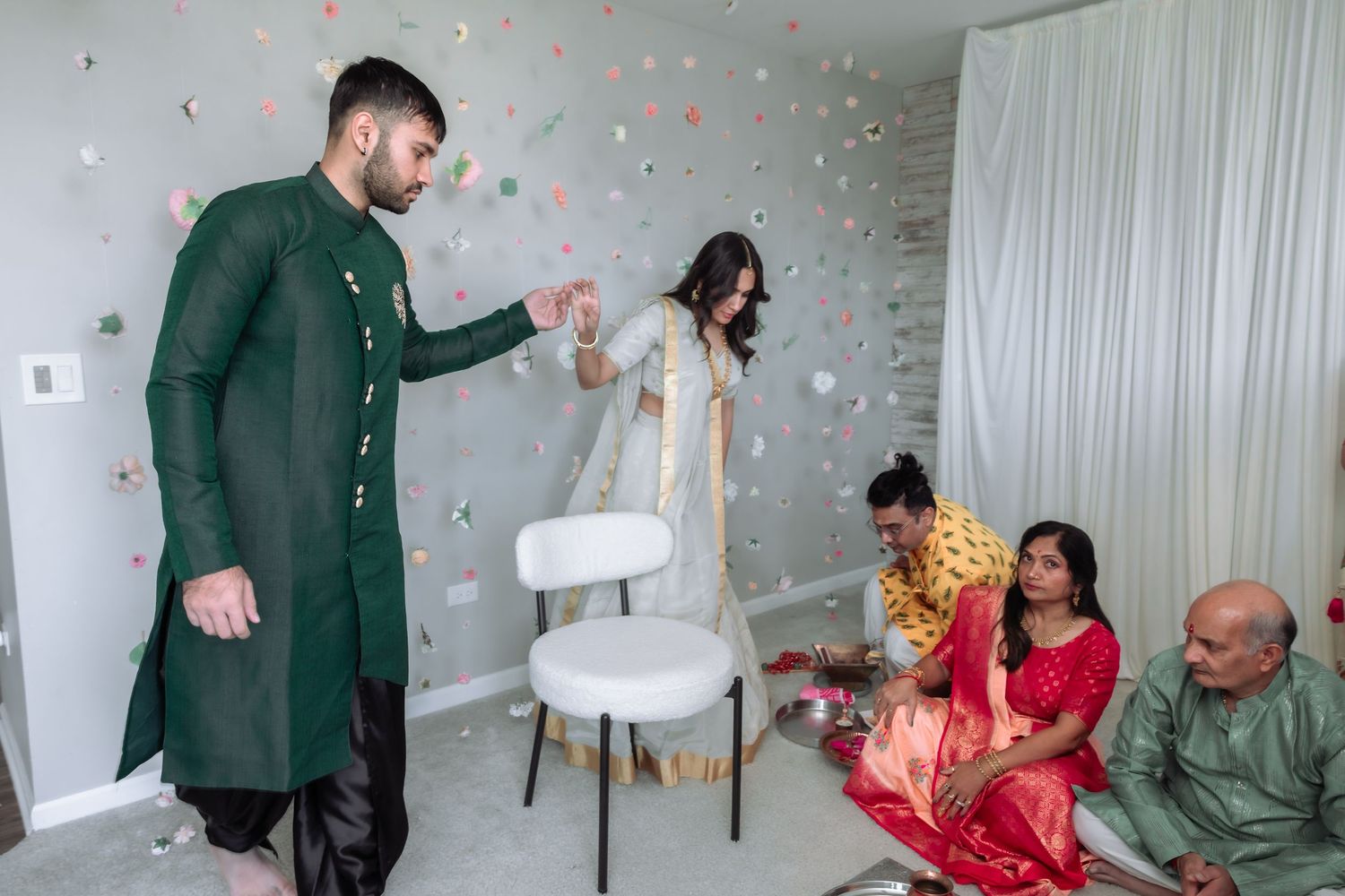 Groom in green sherwani leads bride through room during traditional Indian wedding ceremony with family.
