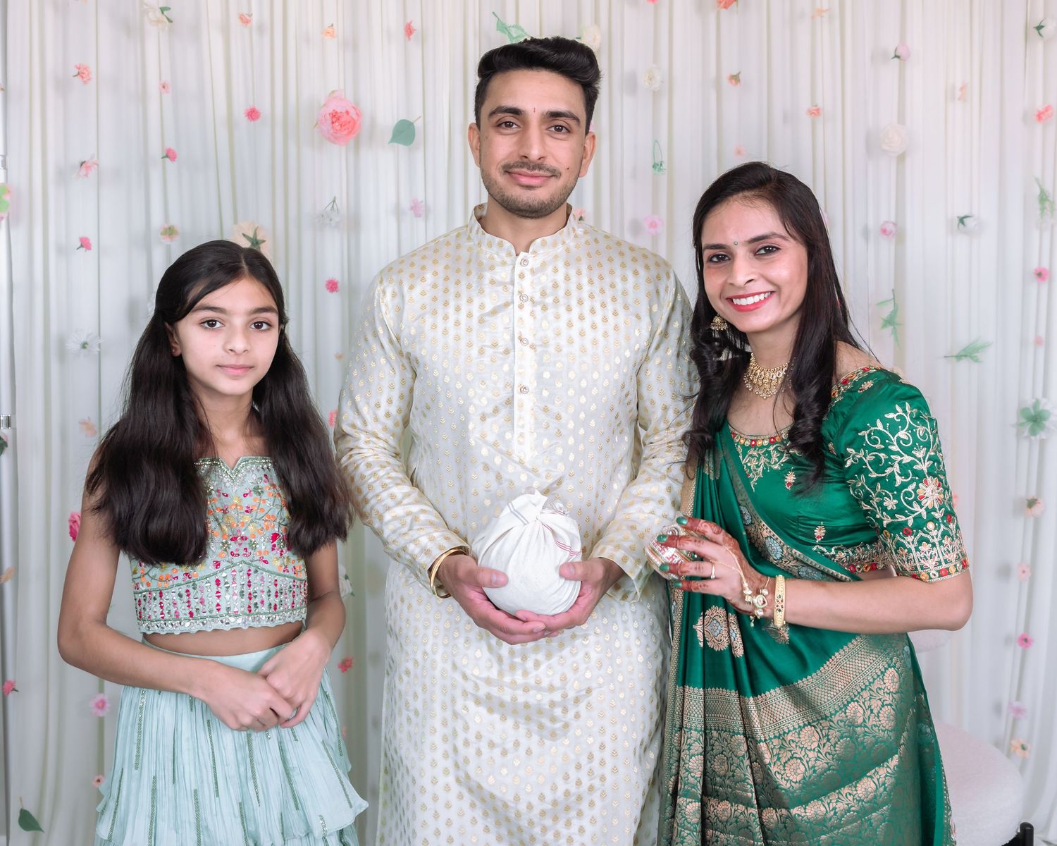 South Asian family of three smiling in traditional Indian clothing at decorated festive celebration.
