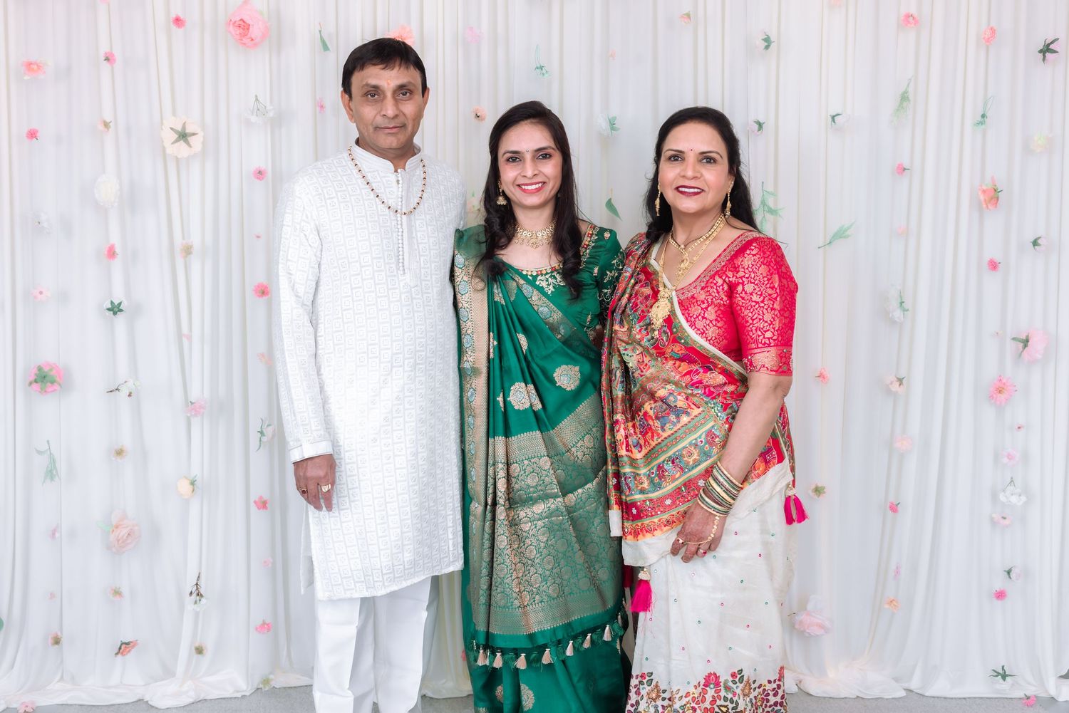 Three people in traditional Indian clothing pose together, two women in green and pink sarees with a man in white kurta.