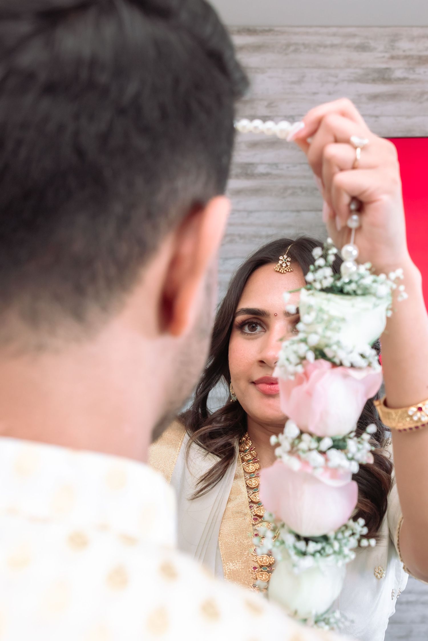 Indian bride in white lehenga holding floral garland with pink roses during traditional engagement ceremony.