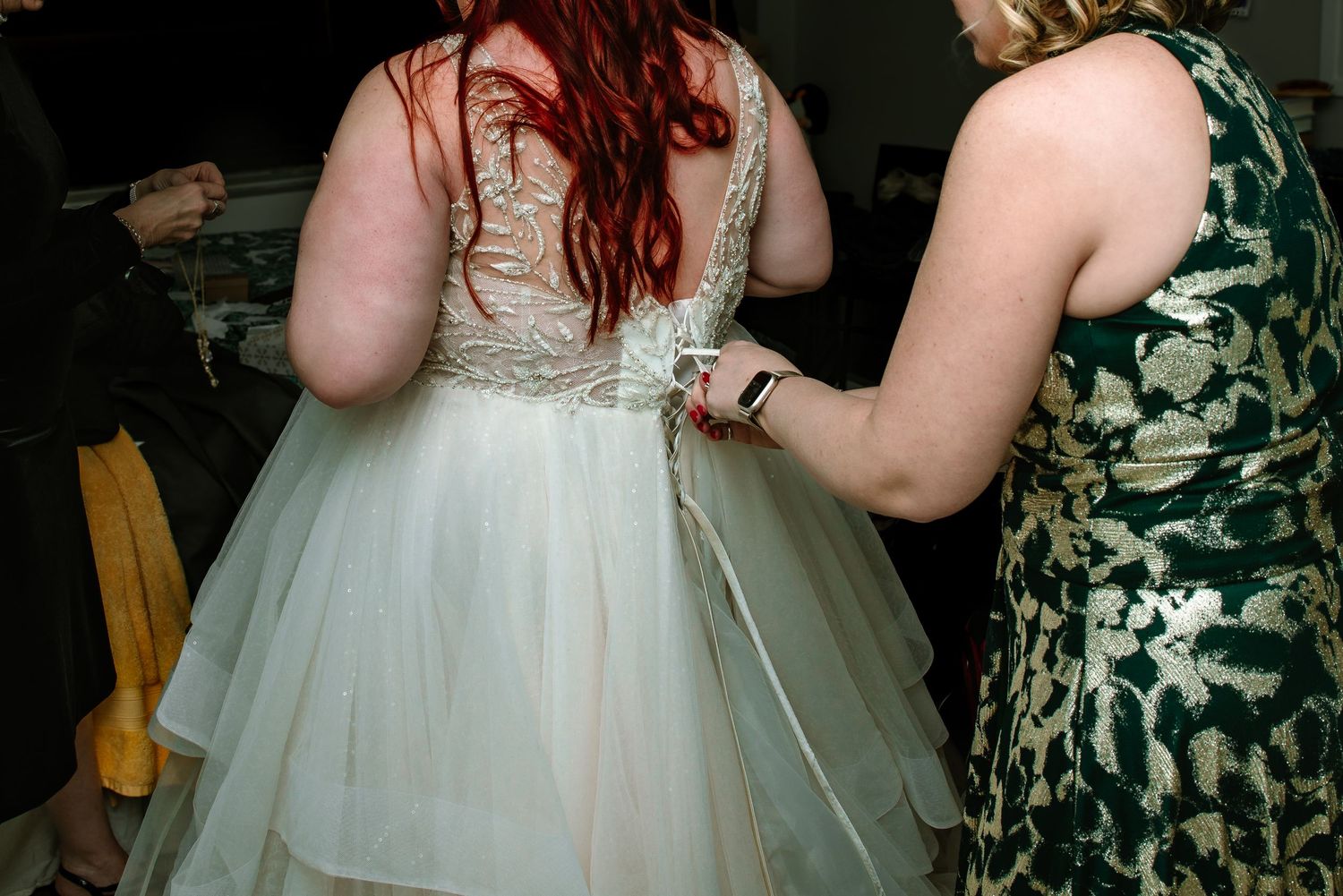Bride in white wedding gown getting dress laced up by bridesmaid in green floral dress.