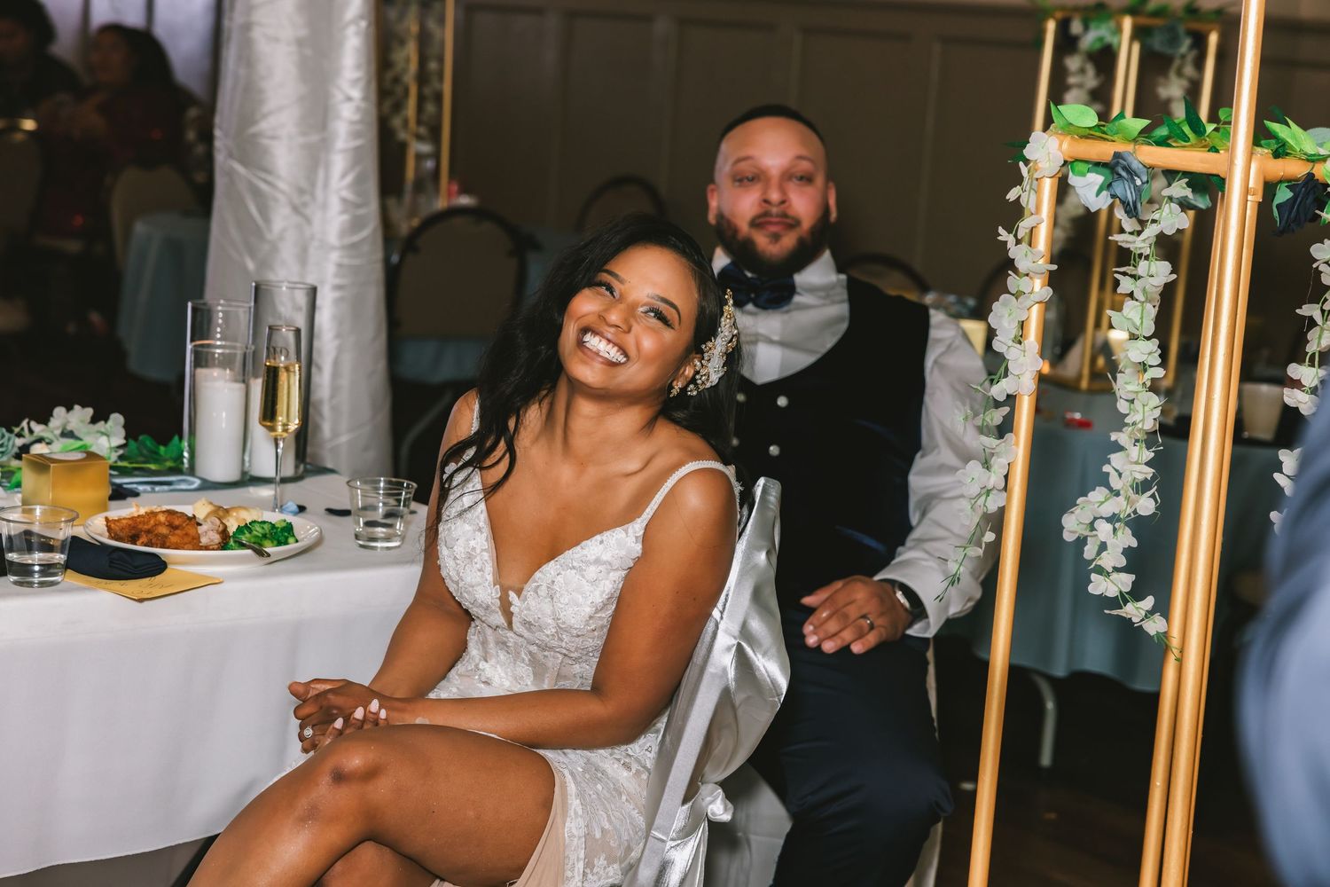 Bride laughing joyfully while seated at wedding reception table with groom behind her.