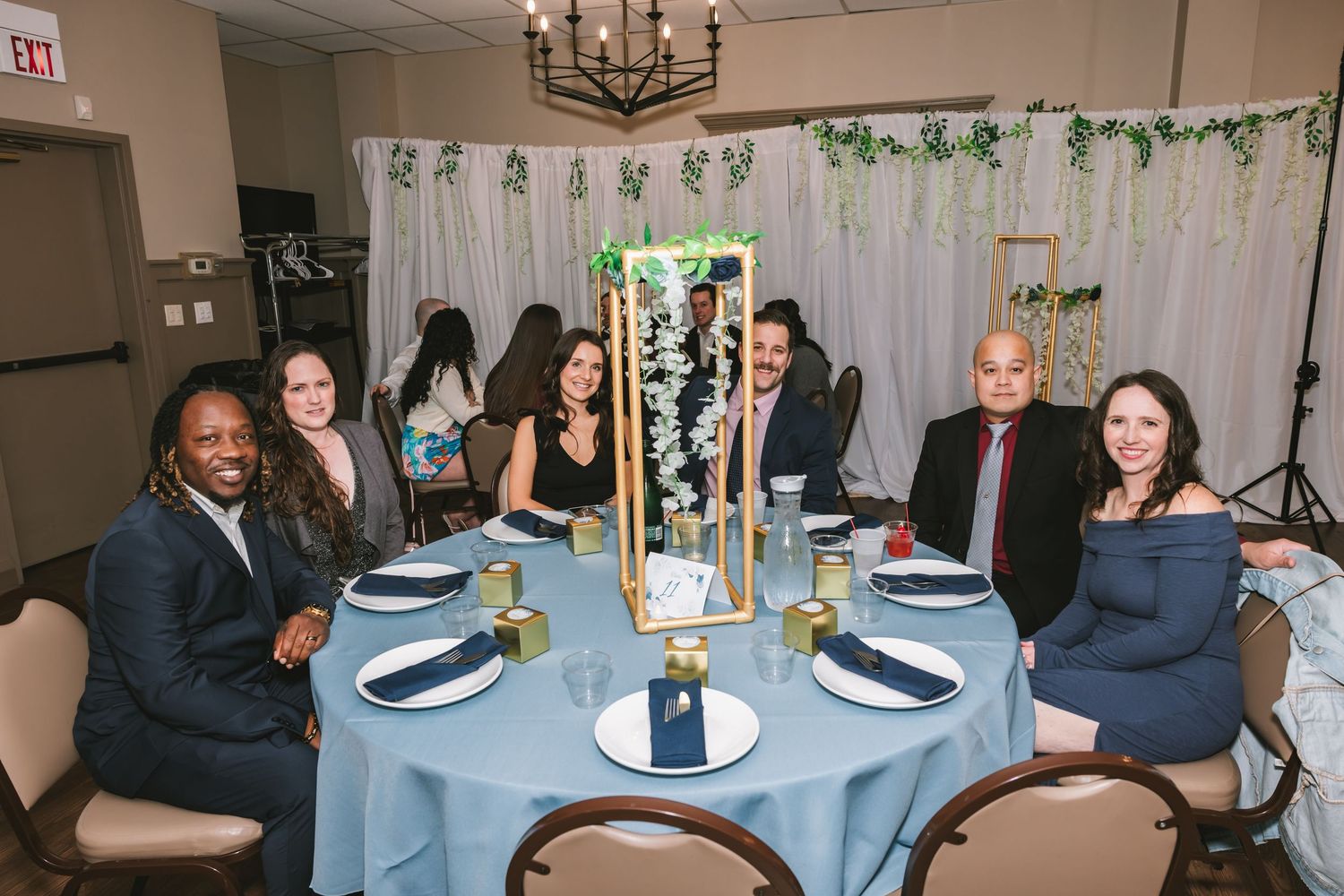 Six wedding guests smiling at decorated reception table with blue linens and tall floral centerpiece.