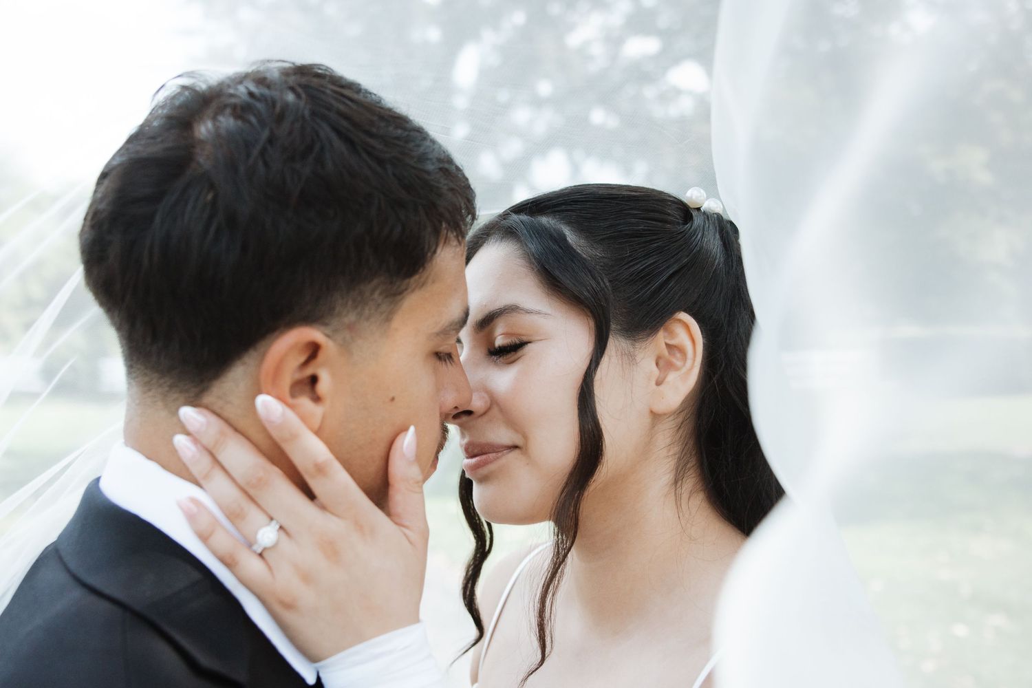 Bride and groom nearly kissing under a white wedding veil, bride wearing diamond ring and white wedding dress.