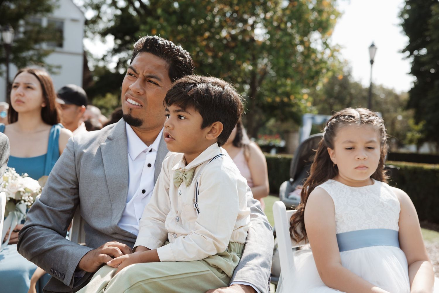 A man in a gray suit sits with two children dressed in formal attire at an outdoor wedding ceremony.