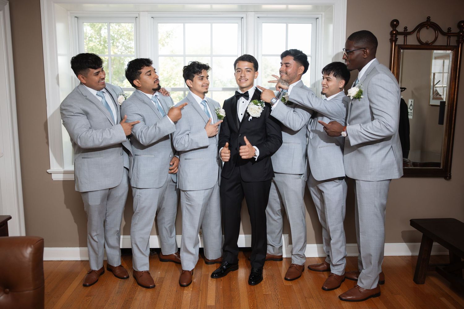 Groom in black tuxedo surrounded by six groomsmen in light gray suits giving thumbs up indoors.