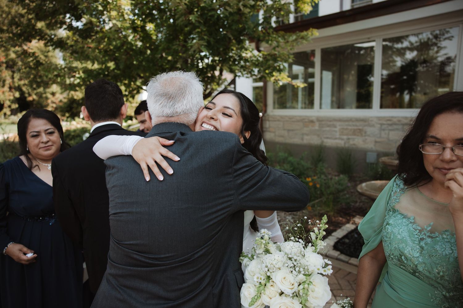 Bride in white gown embracing an older gentleman guest while holding white flower bouquet outdoors.