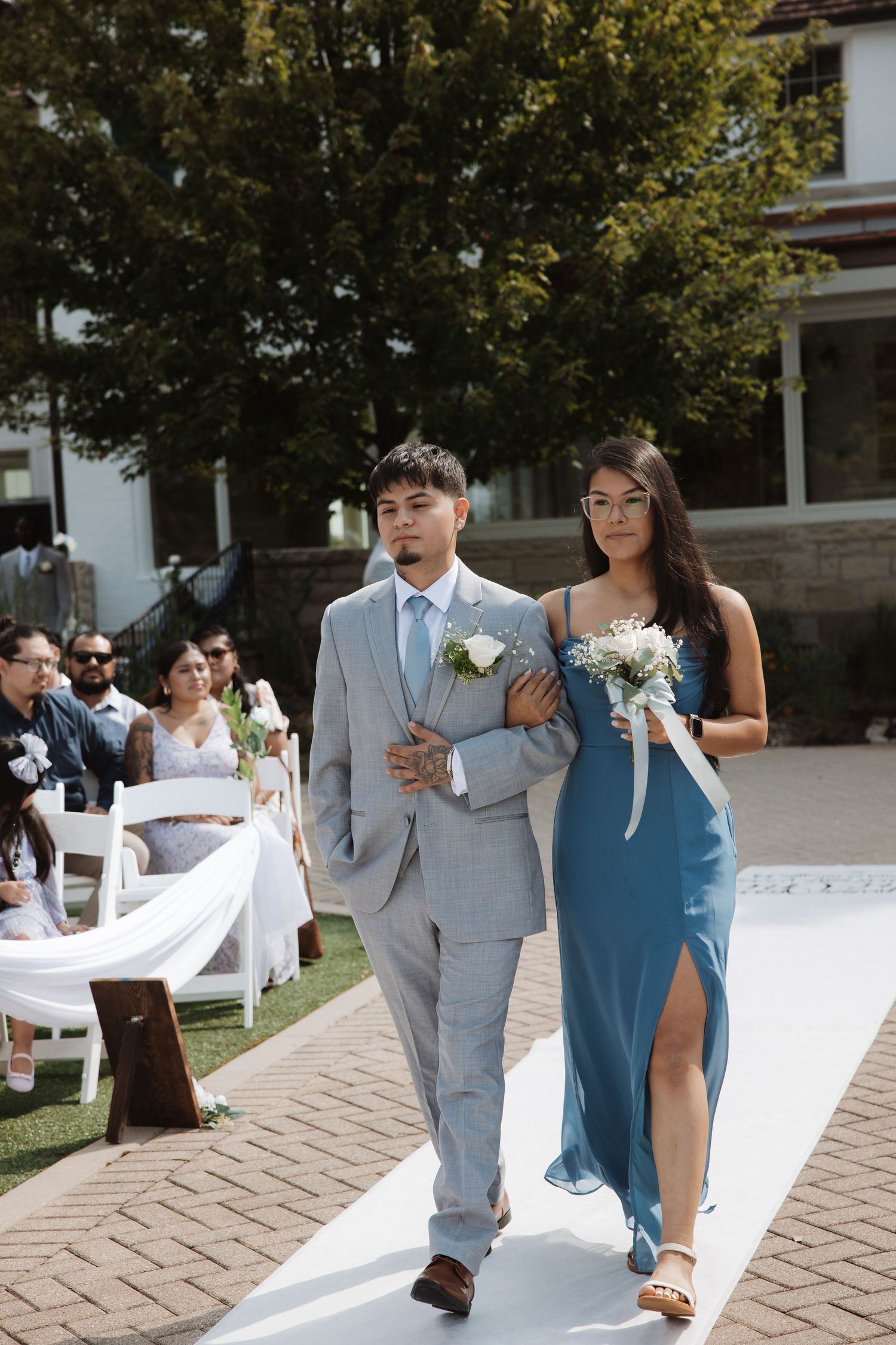 Groomsman in gray suit escorts bridesmaid in blue dress down outdoor wedding aisle.