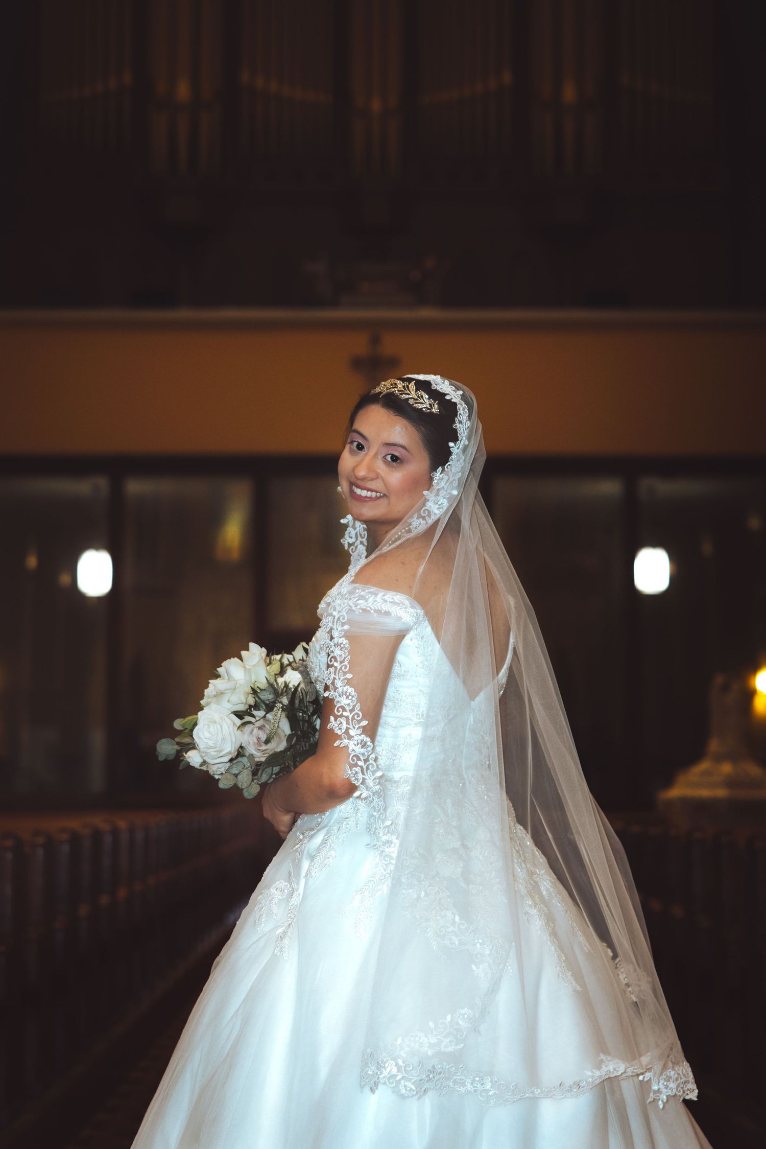 Smiling bride in lace wedding gown and tiara holds white bouquet inside ornate church interior.