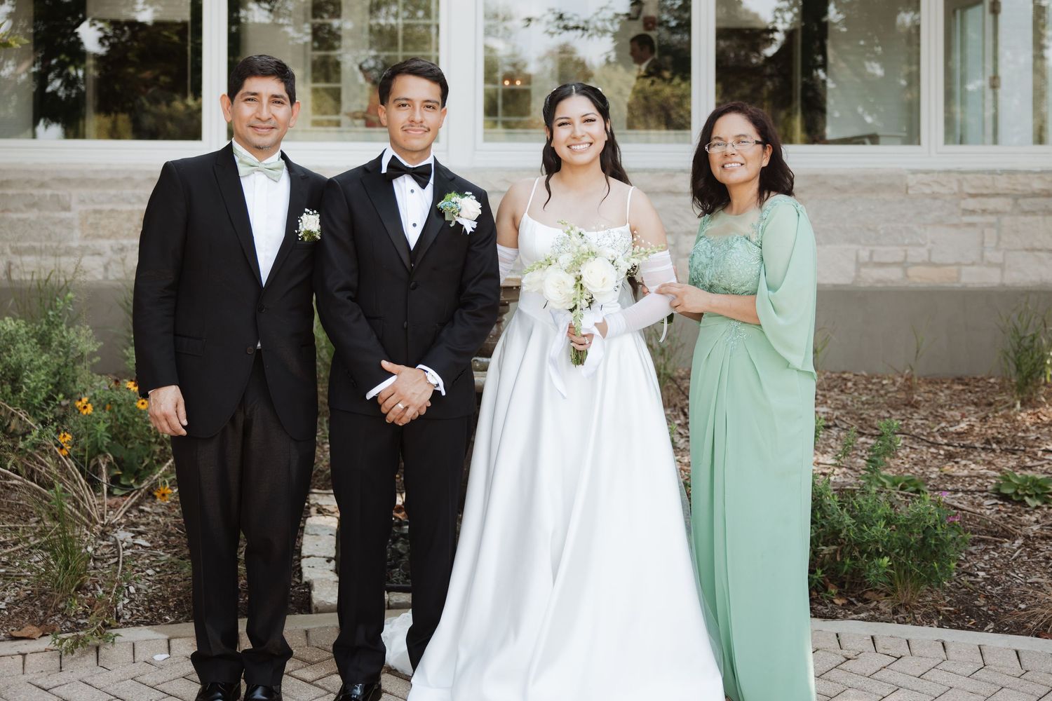Bride and groom pose with two family members outside a wedding venue, bride in white gown holding bouquet.