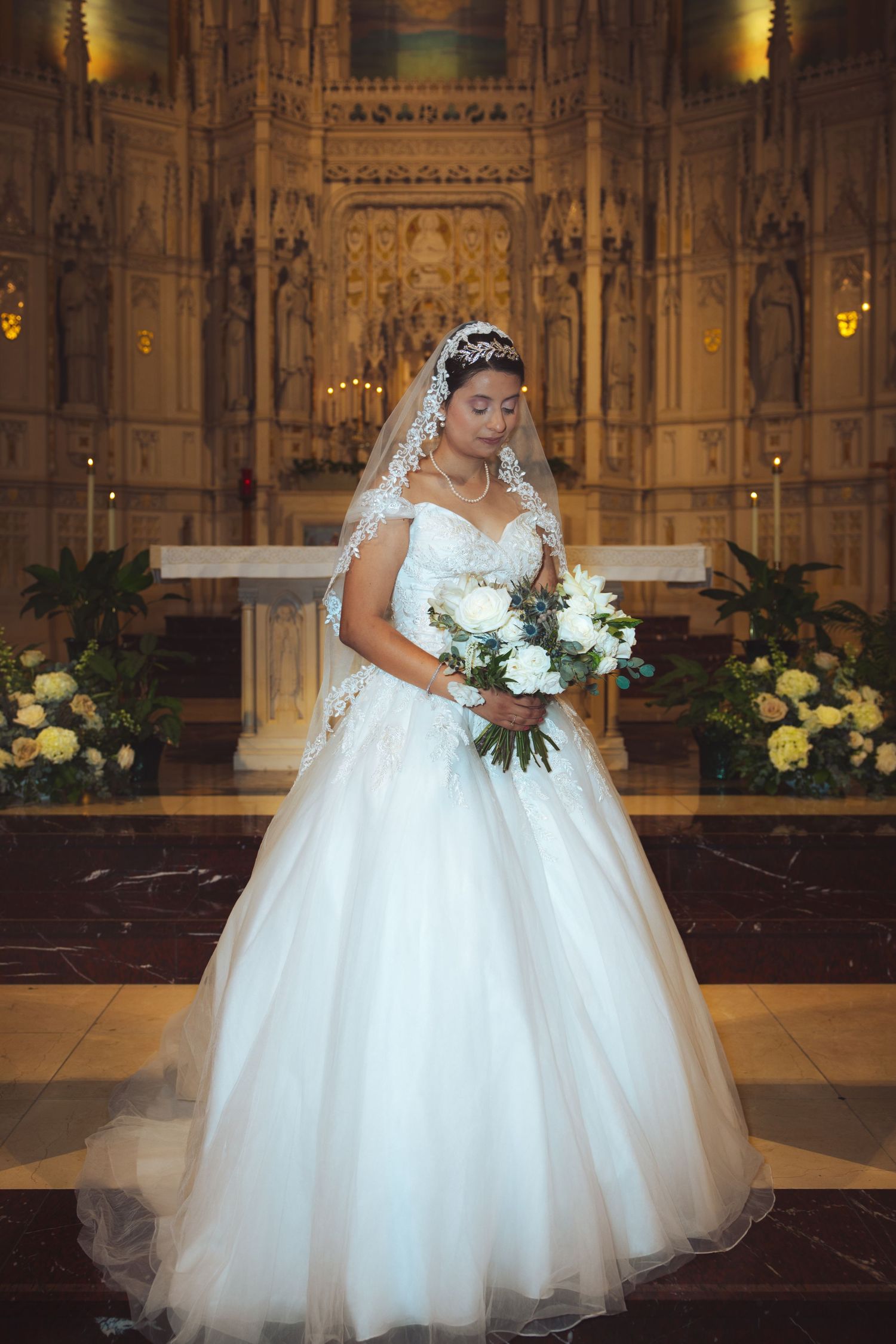 Bride in white off-shoulder ball gown holding white bouquet standing before golden Gothic church altar.