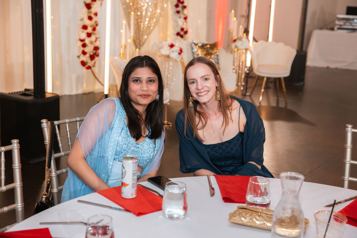 Two women smile together at decorated table during Indian wedding reception dinner.
