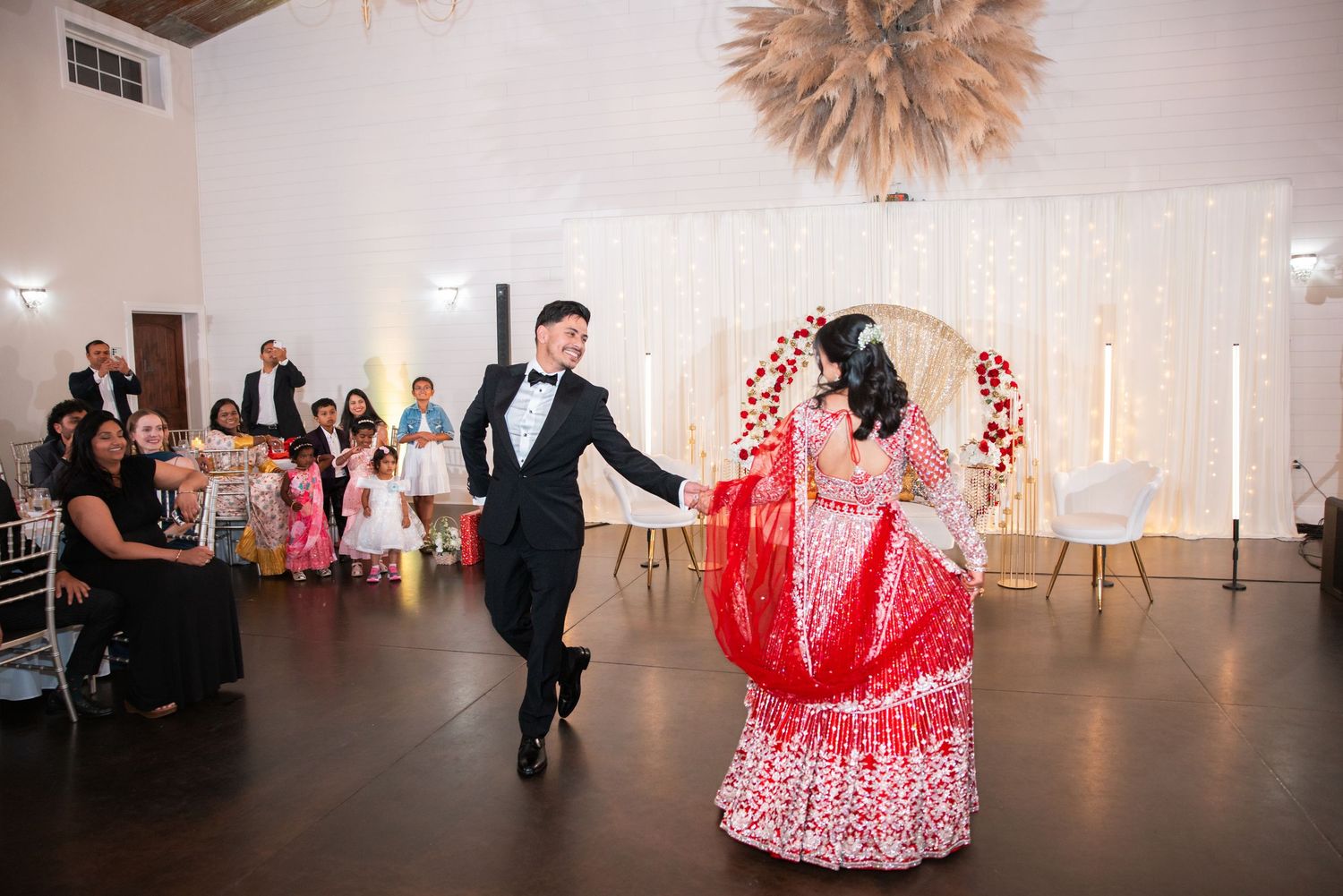 Groom in tuxedo and bride in vibrant red lehenga dancing joyfully before guests at Indian wedding reception.