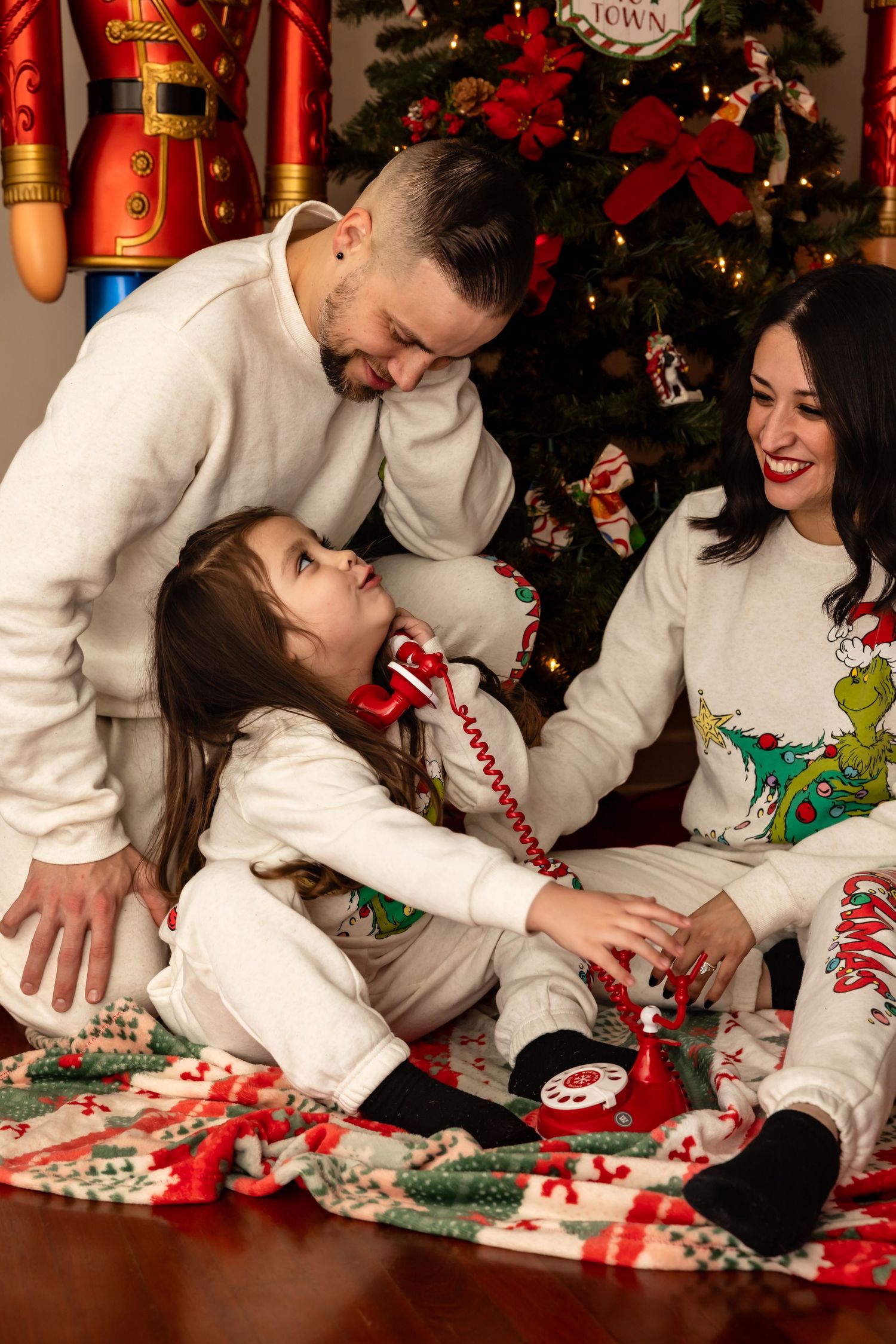 Family of three in matching Grinch Christmas pajamas sitting together near a festively decorated holiday tree.
