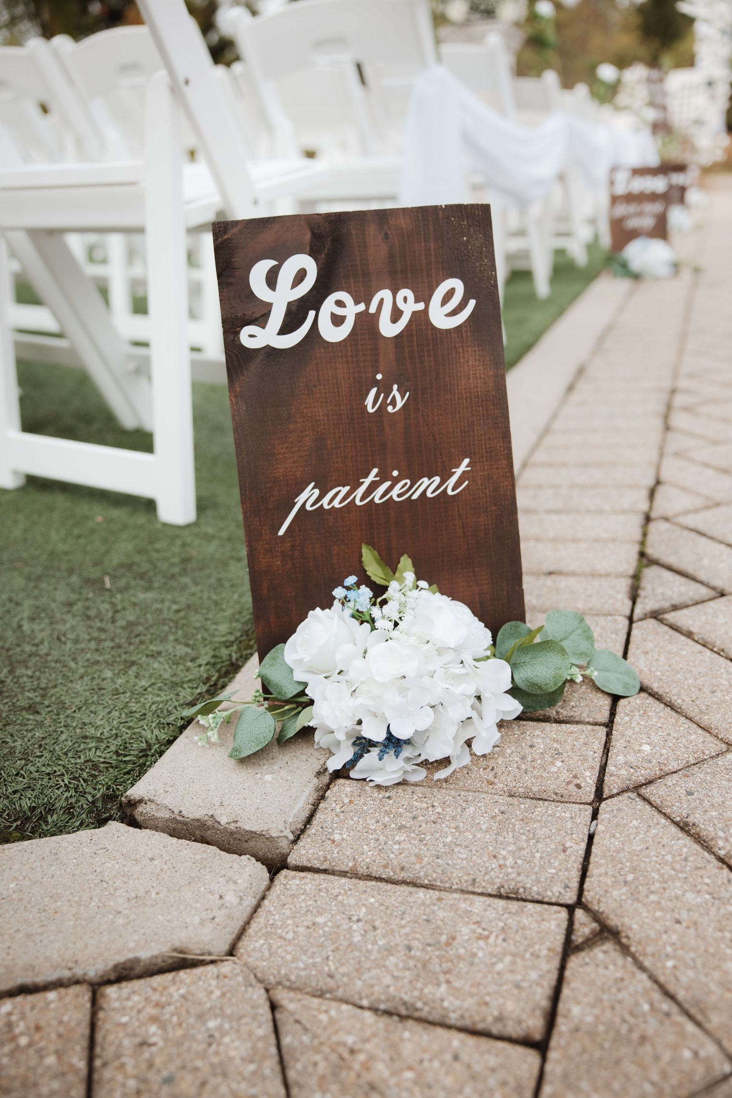 Rustic wooden wedding sign reading Love is patient with white hydrangeas along a brick ceremony aisle.