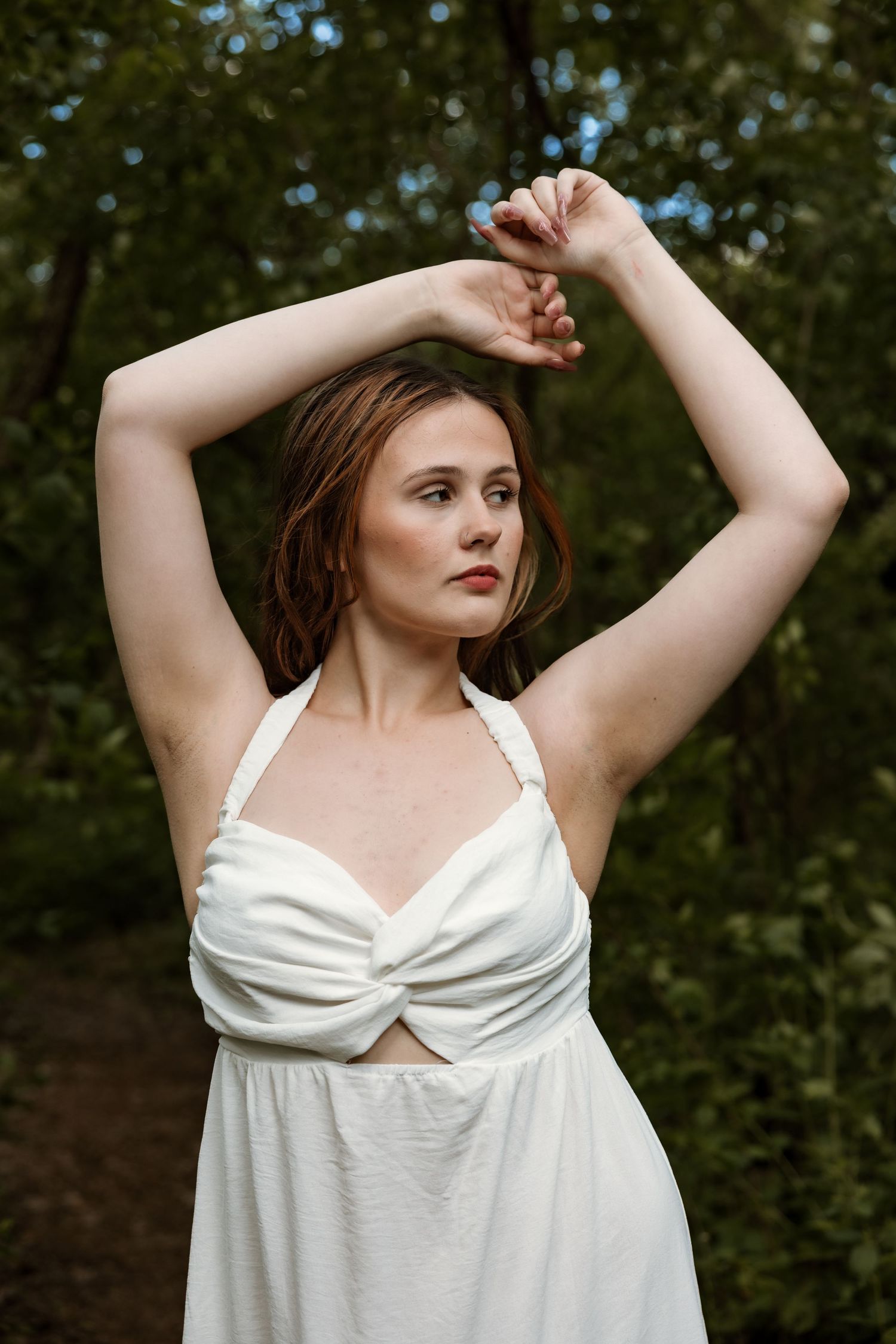 Redhead woman in white halter dress raises arms gracefully in outdoor forest portrait.
