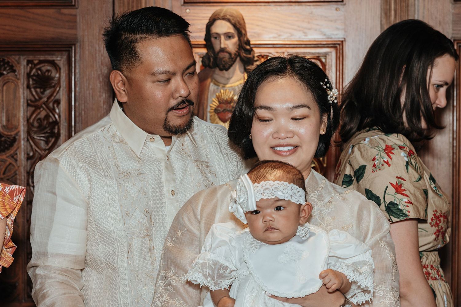 Filipino parents in traditional barong attire proudly hold their baby during a Catholic baptism ceremony.