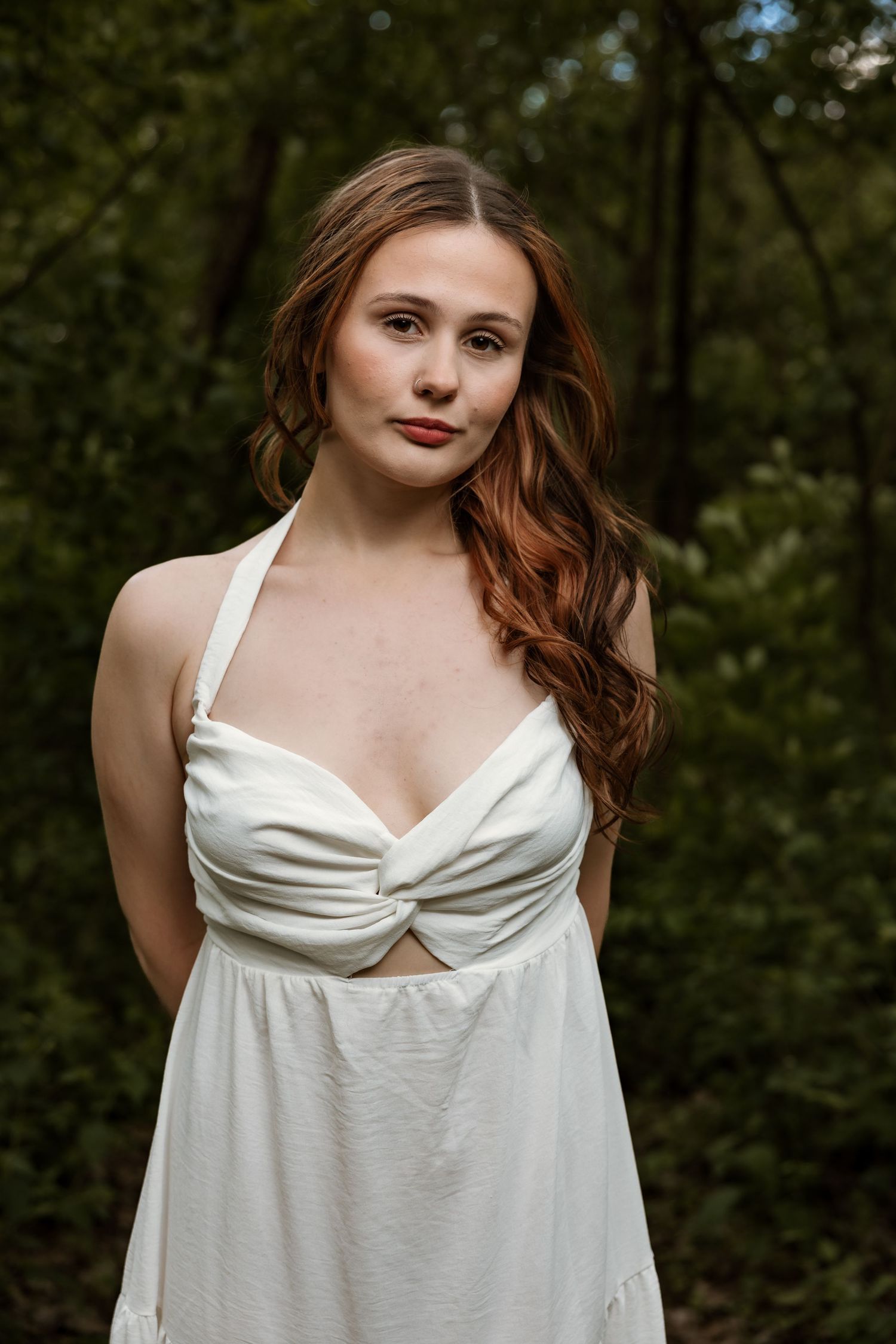 Young woman with auburn hair in white halter dress poses in lush green forest setting.