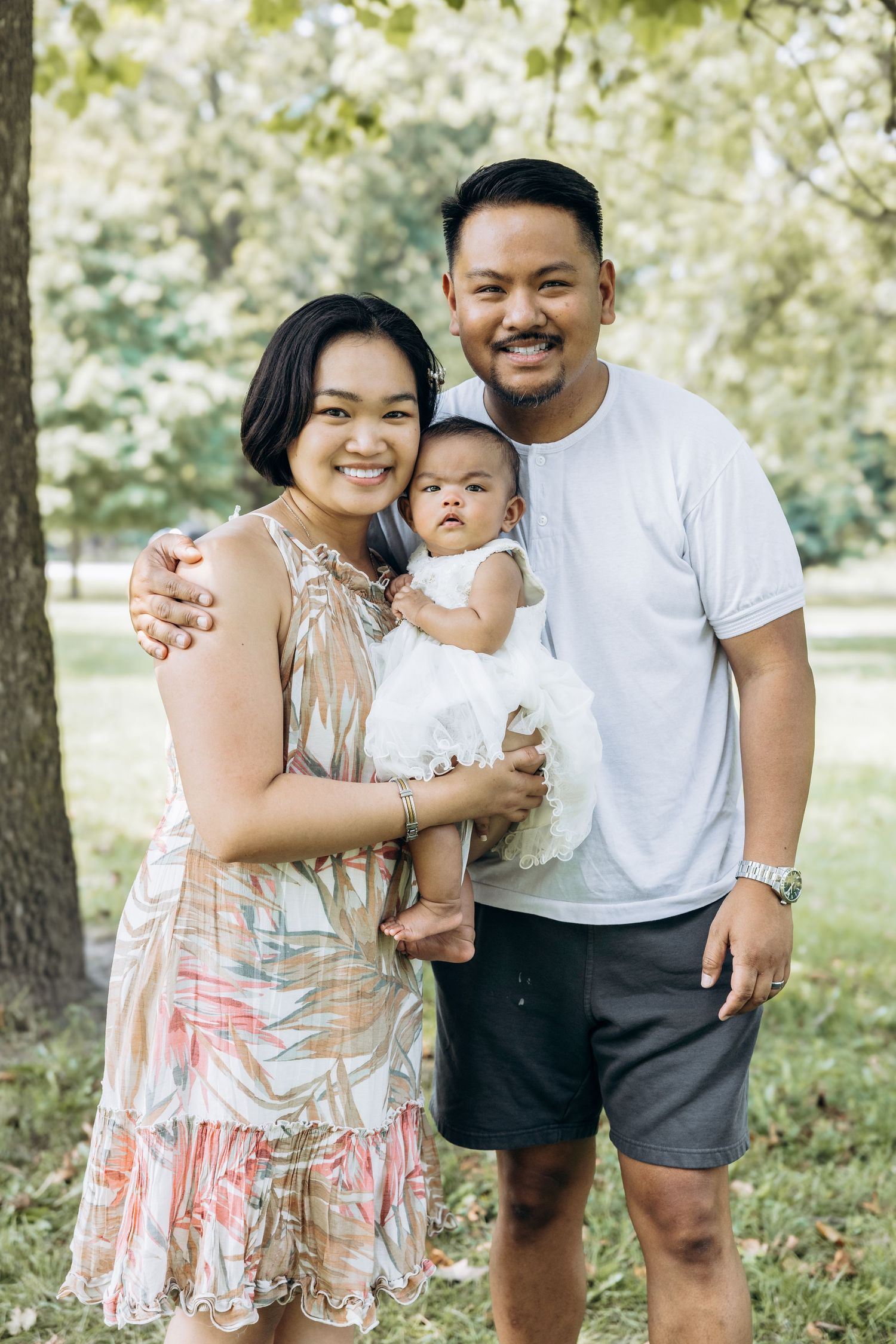 Young Asian family smiles together in park with baby dressed in white.