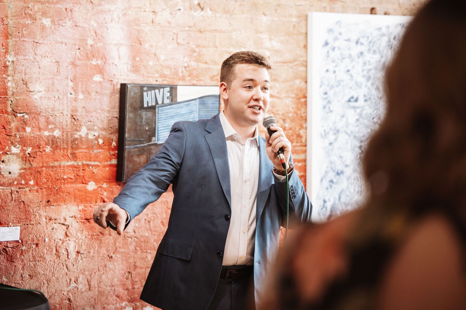 Man in blue blazer speaks into microphone at an indoor event with exposed brick walls.