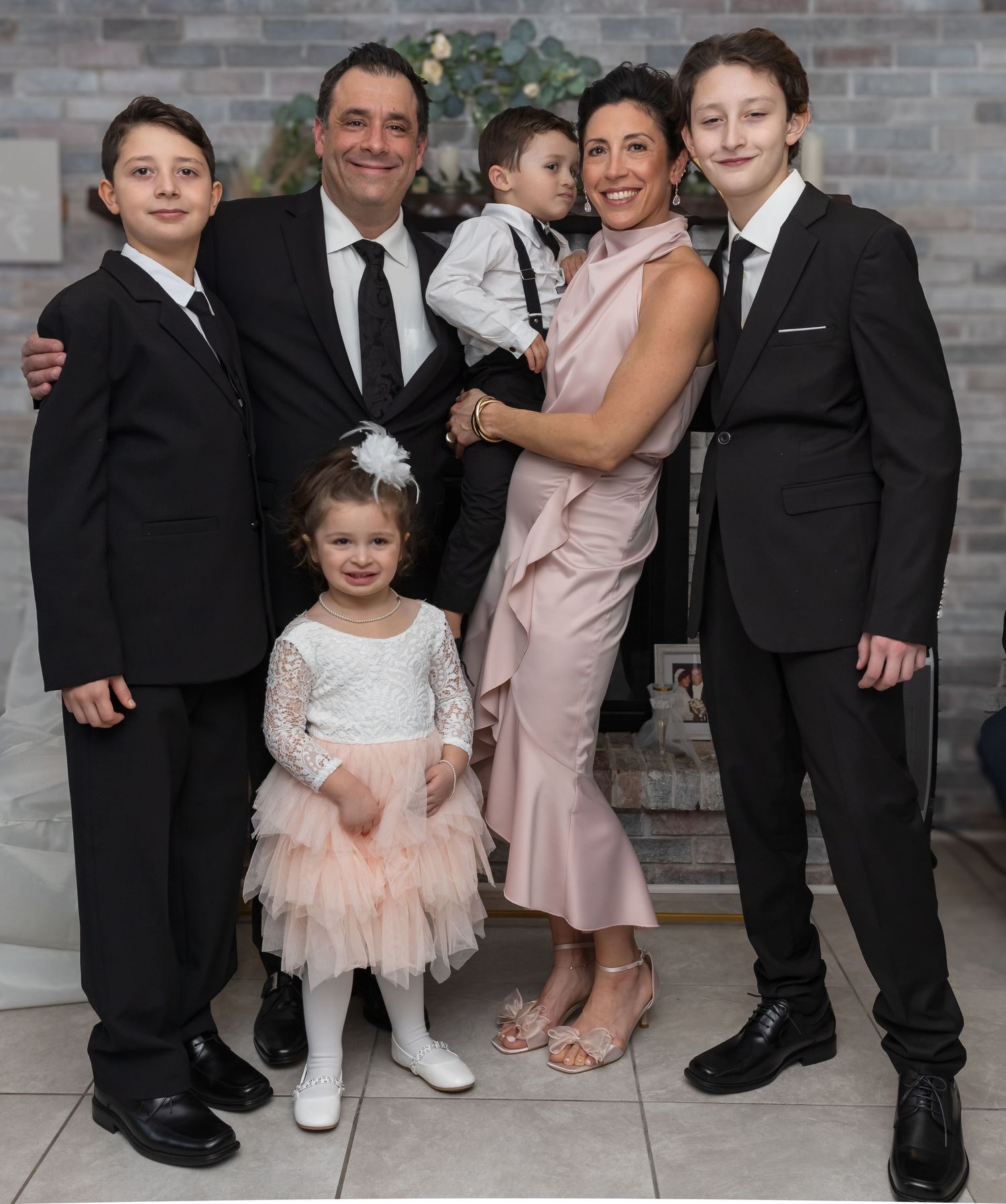 Happy family of six in formal attire posing together at a wedding with brick fireplace backdrop.