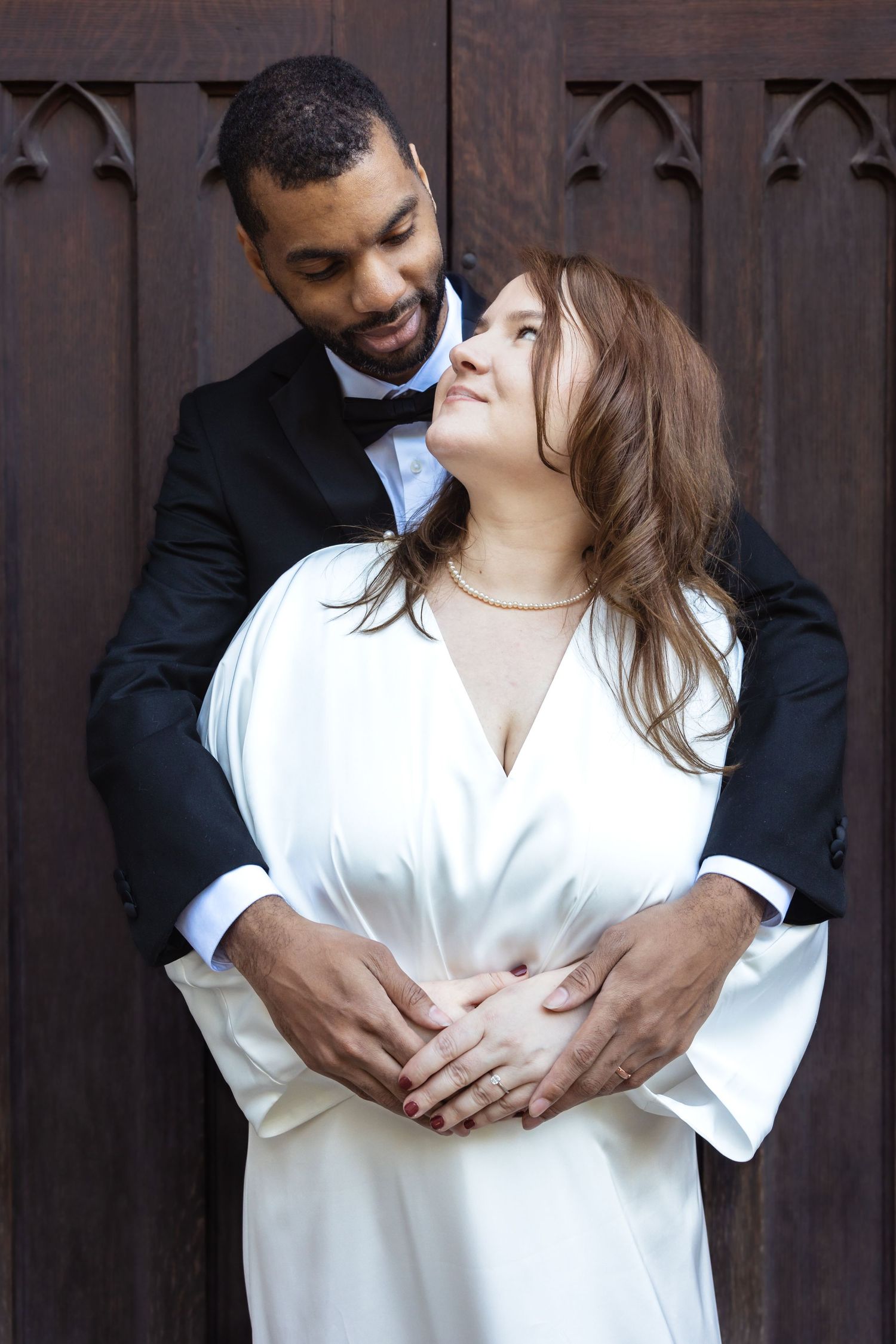 Interracial couple embracing in wedding attire before a wooden church door.
